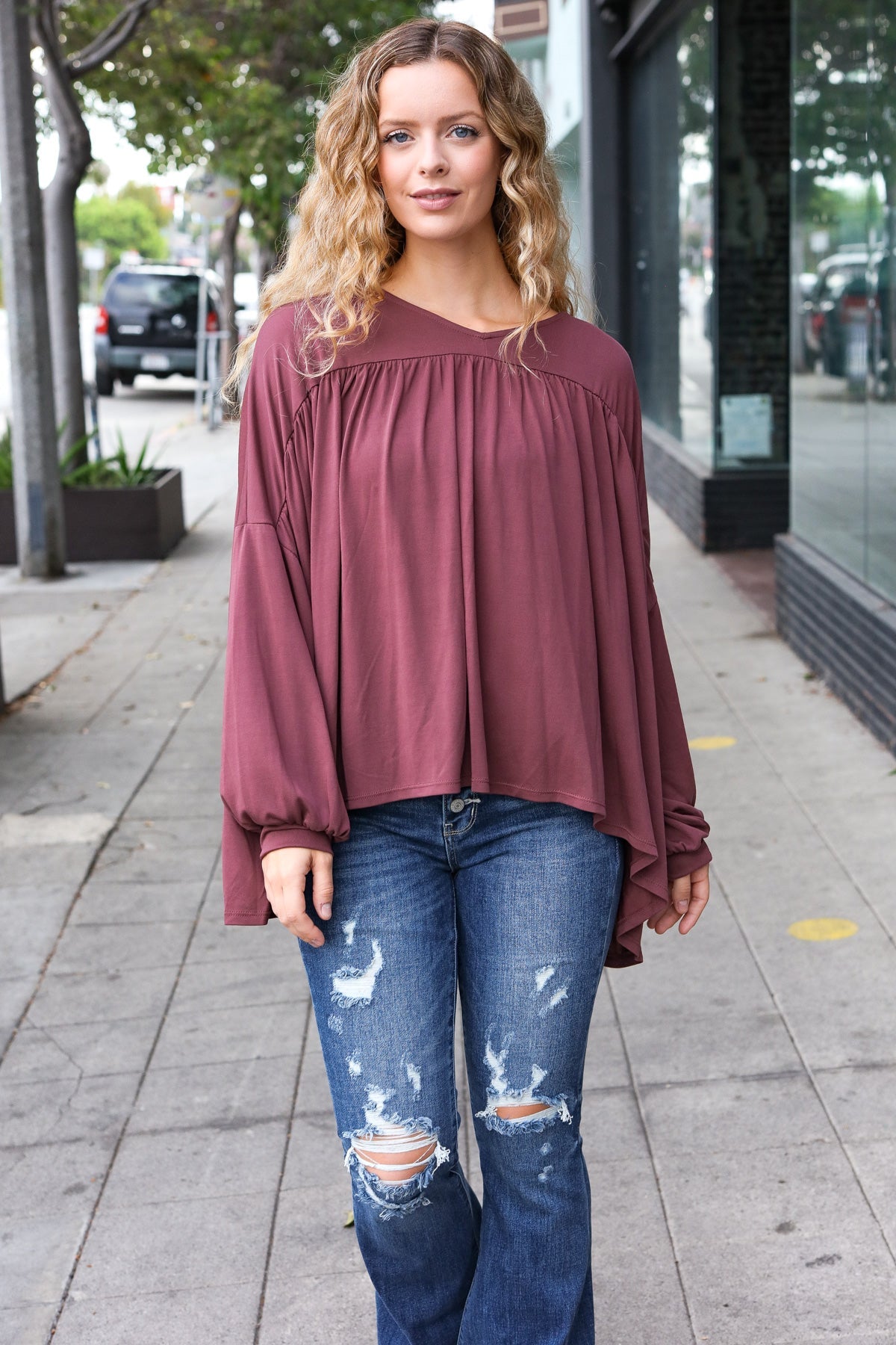 Woman standing outdoors wearing wine In This Groove modal knit top with gathered yoke and loose long sleeves paired with distressed blue jeans