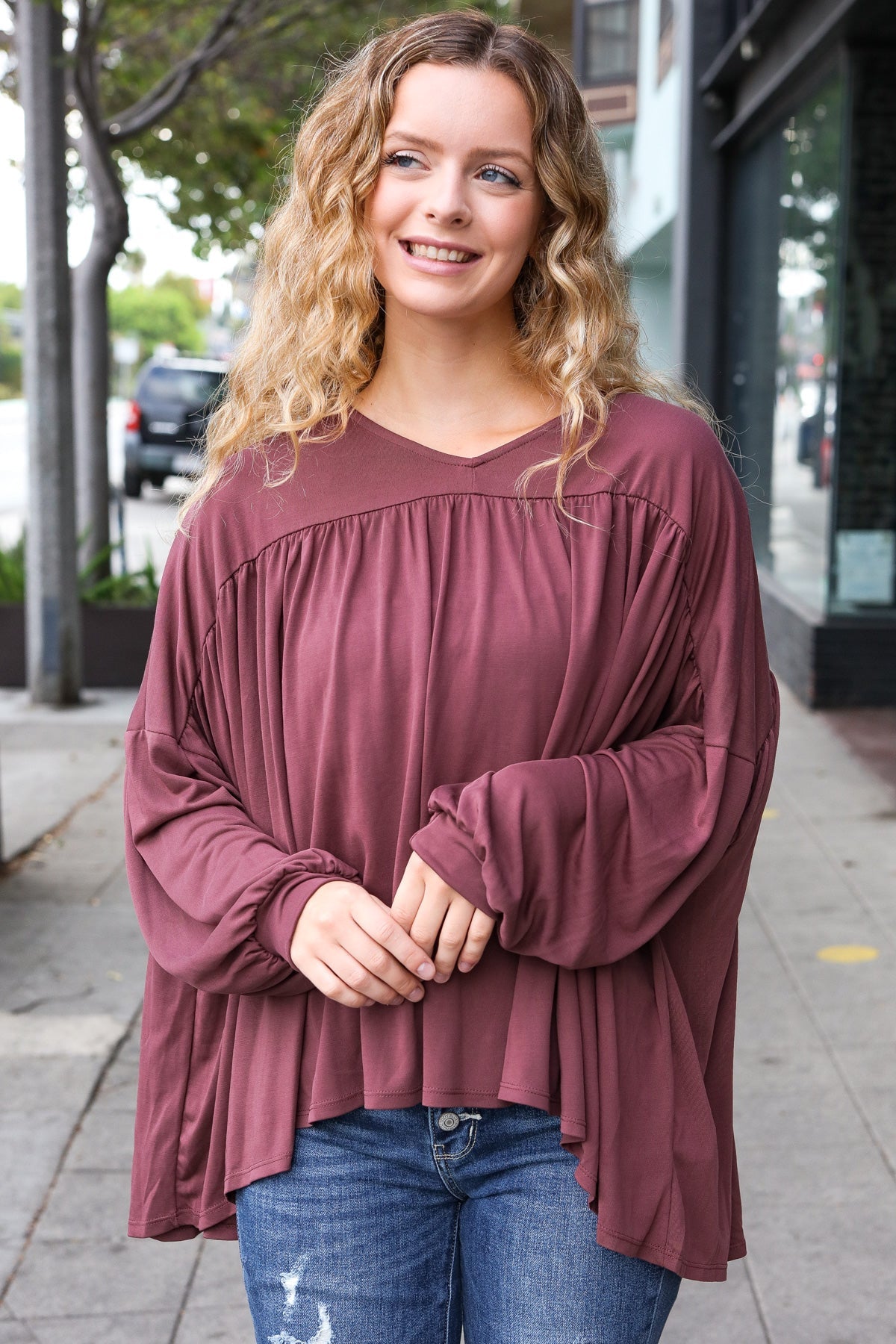 Woman smiling outdoors wearing wine In This Groove modal knit top with gathered yoke and oversized long sleeves paired with blue jeans