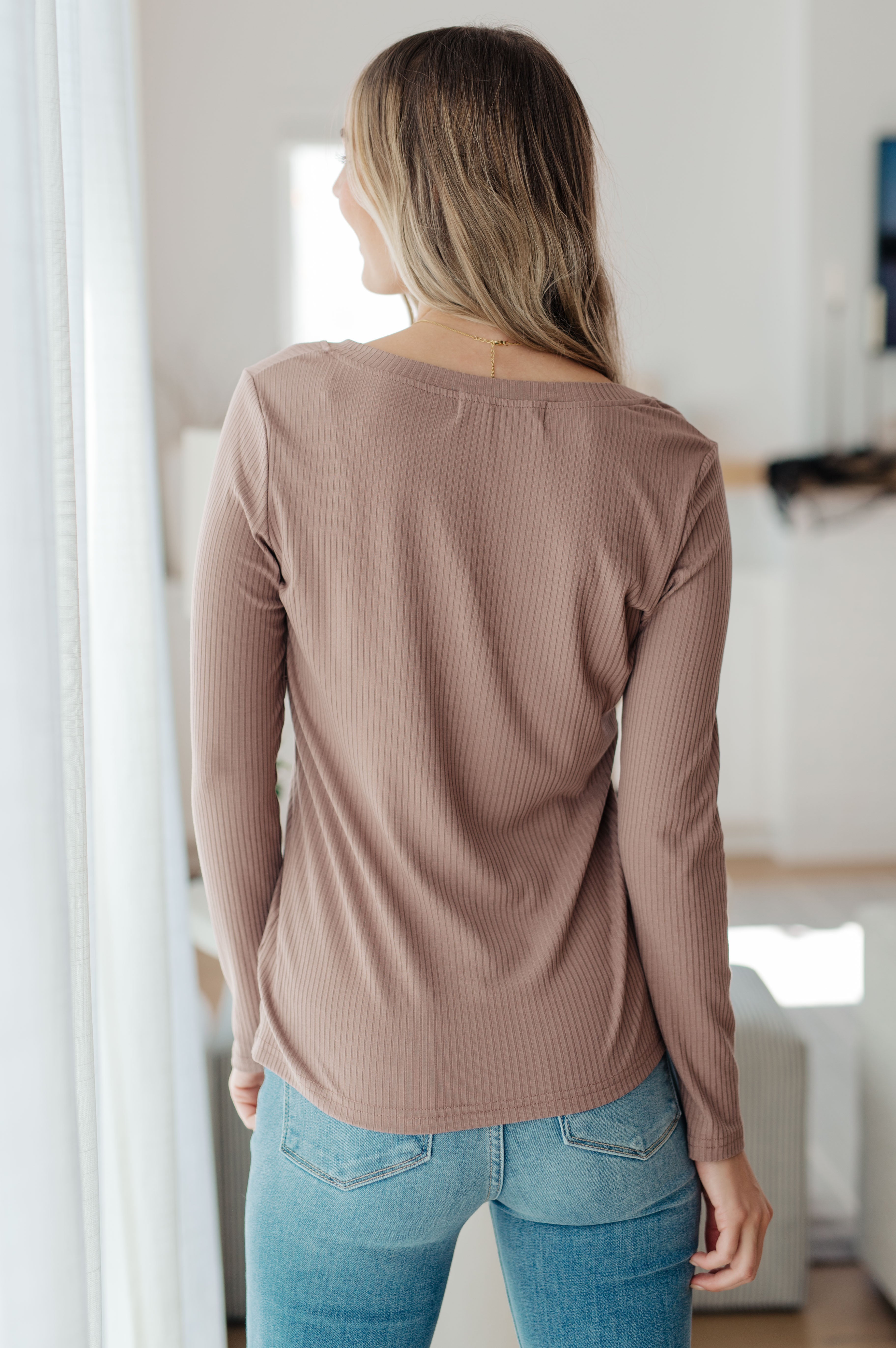 Woman standing indoors wearing taupe Indeed You Do long sleeve V-neck top with textured fabric and light blue jeans viewed from back