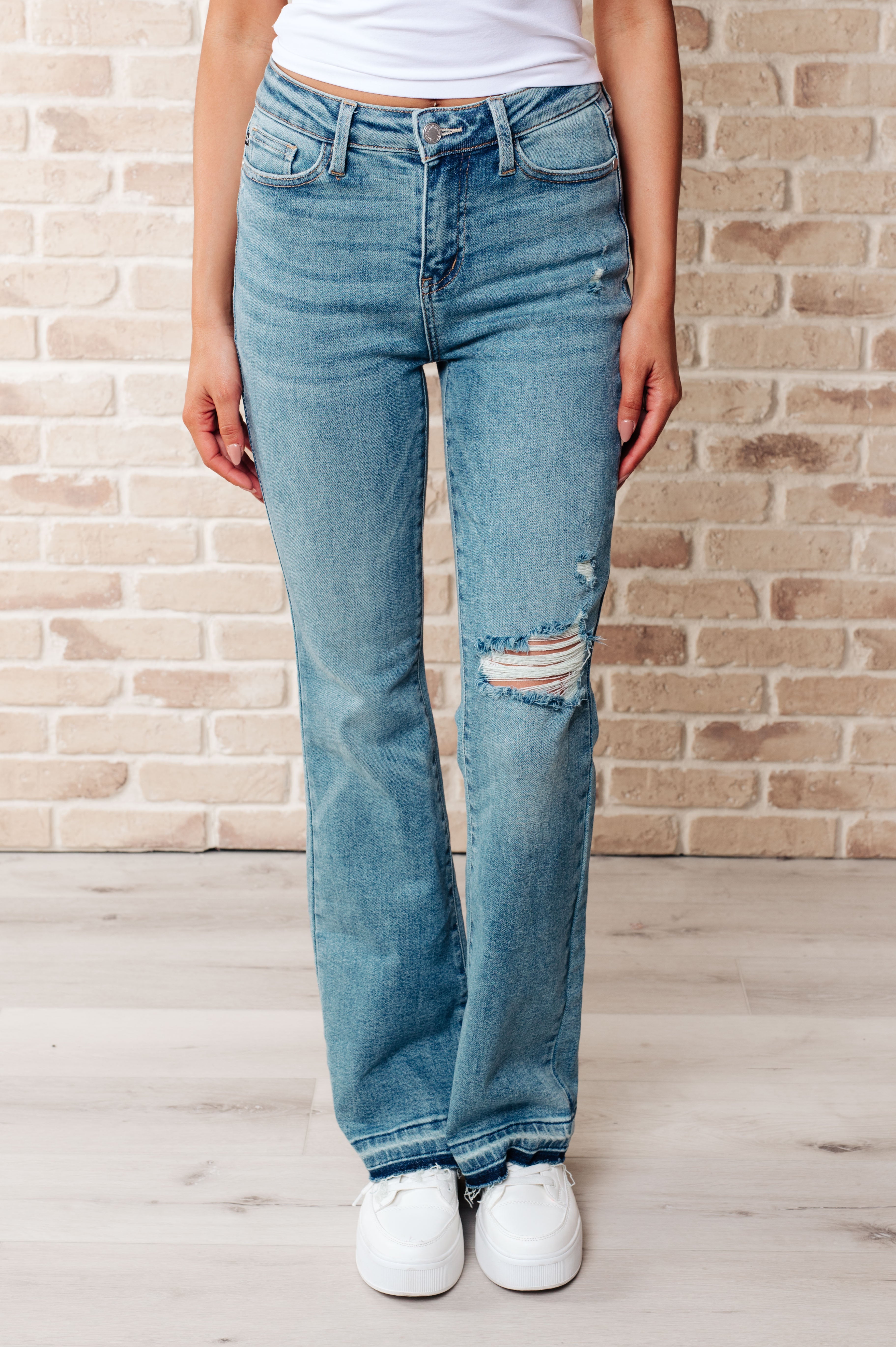 Front view of person wearing blue Isla mid rise bootcut jeans with distressed knee and raw hems paired with white sneakers indoors