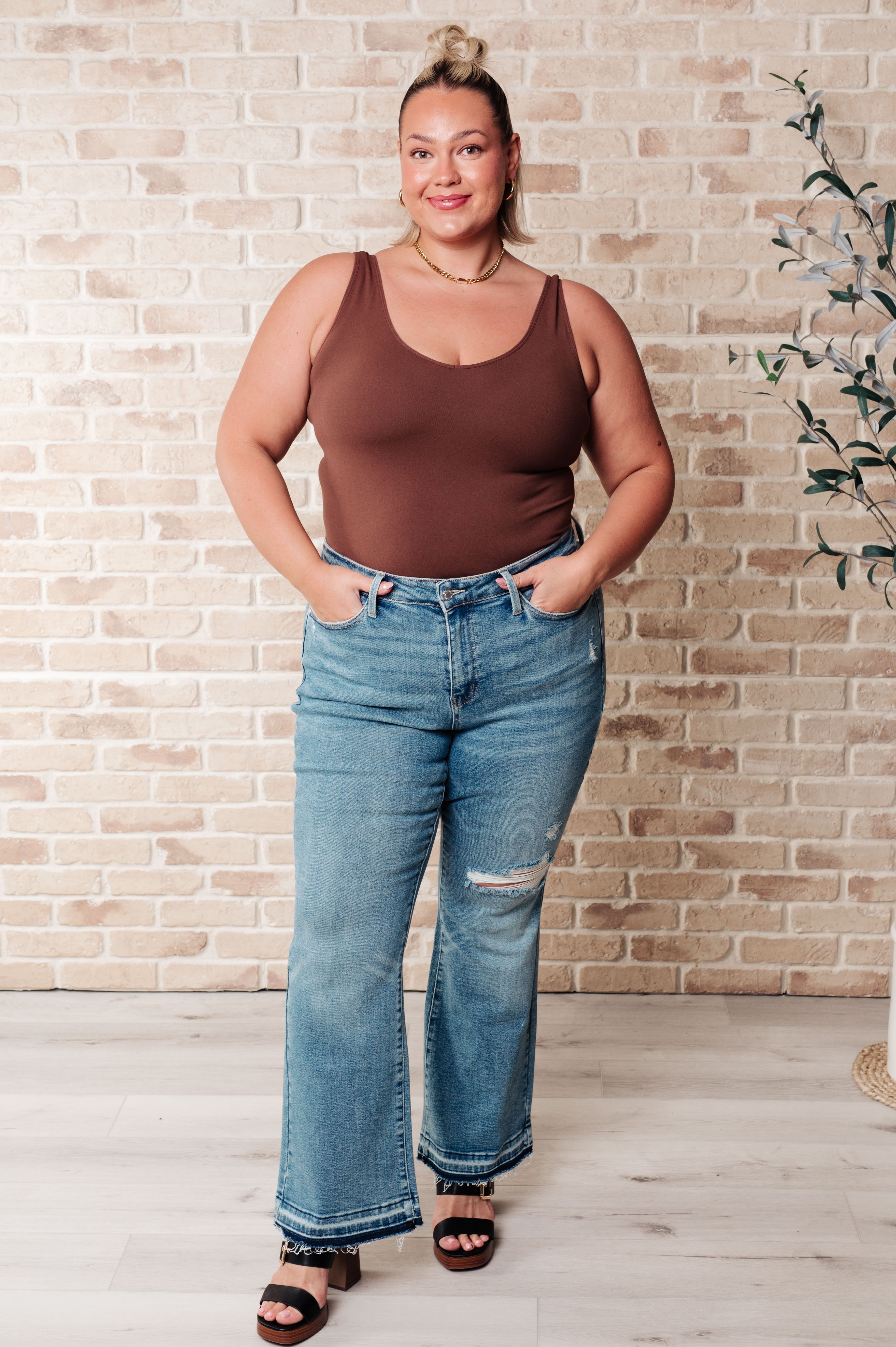 Person wearing blue Isla mid rise bootcut jeans with distressed knee detail and released hems paired with black sandals indoors