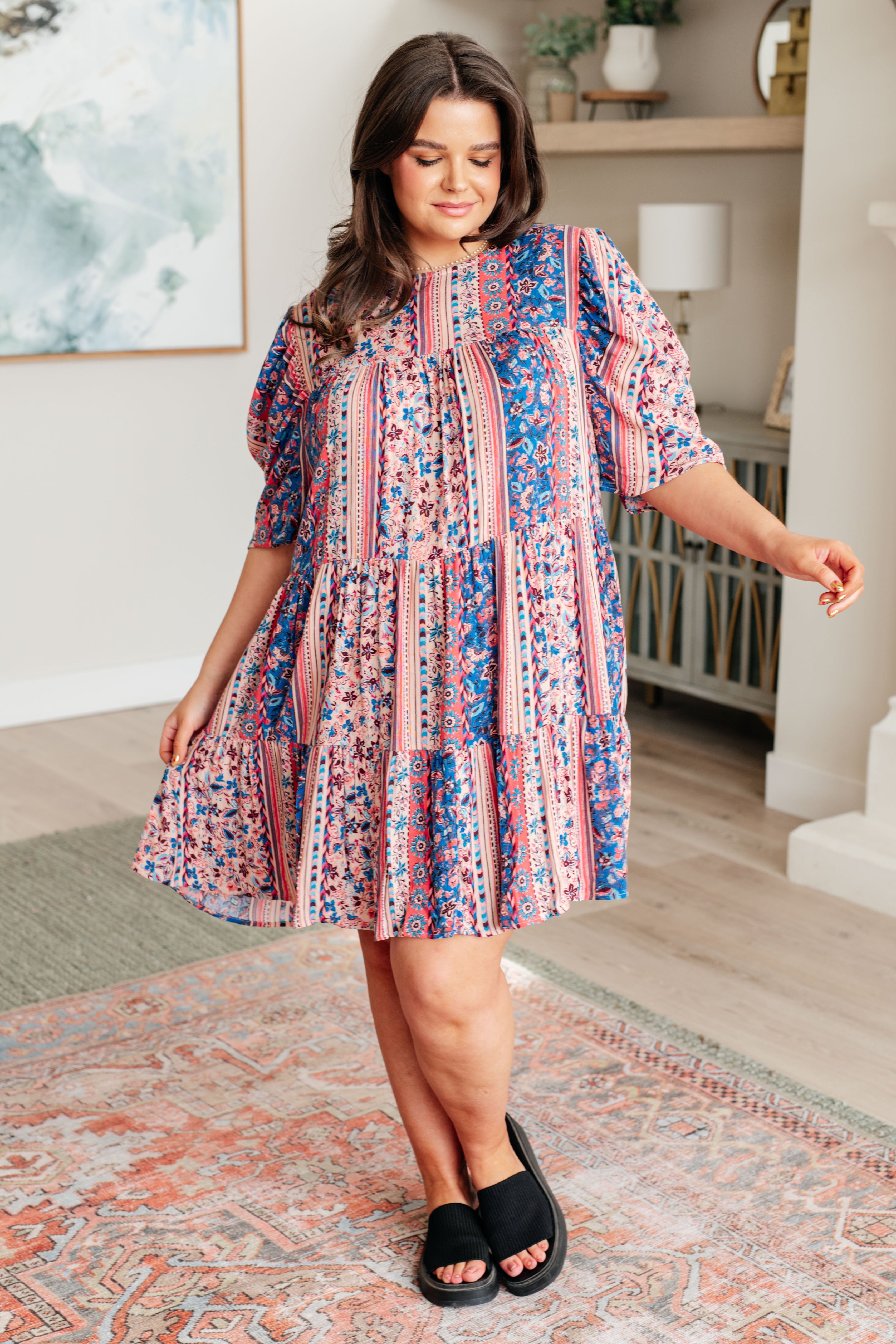 Woman wearing a loose pink and blue patterned tiered dress with elbow-length puff sleeves and black slide sandals