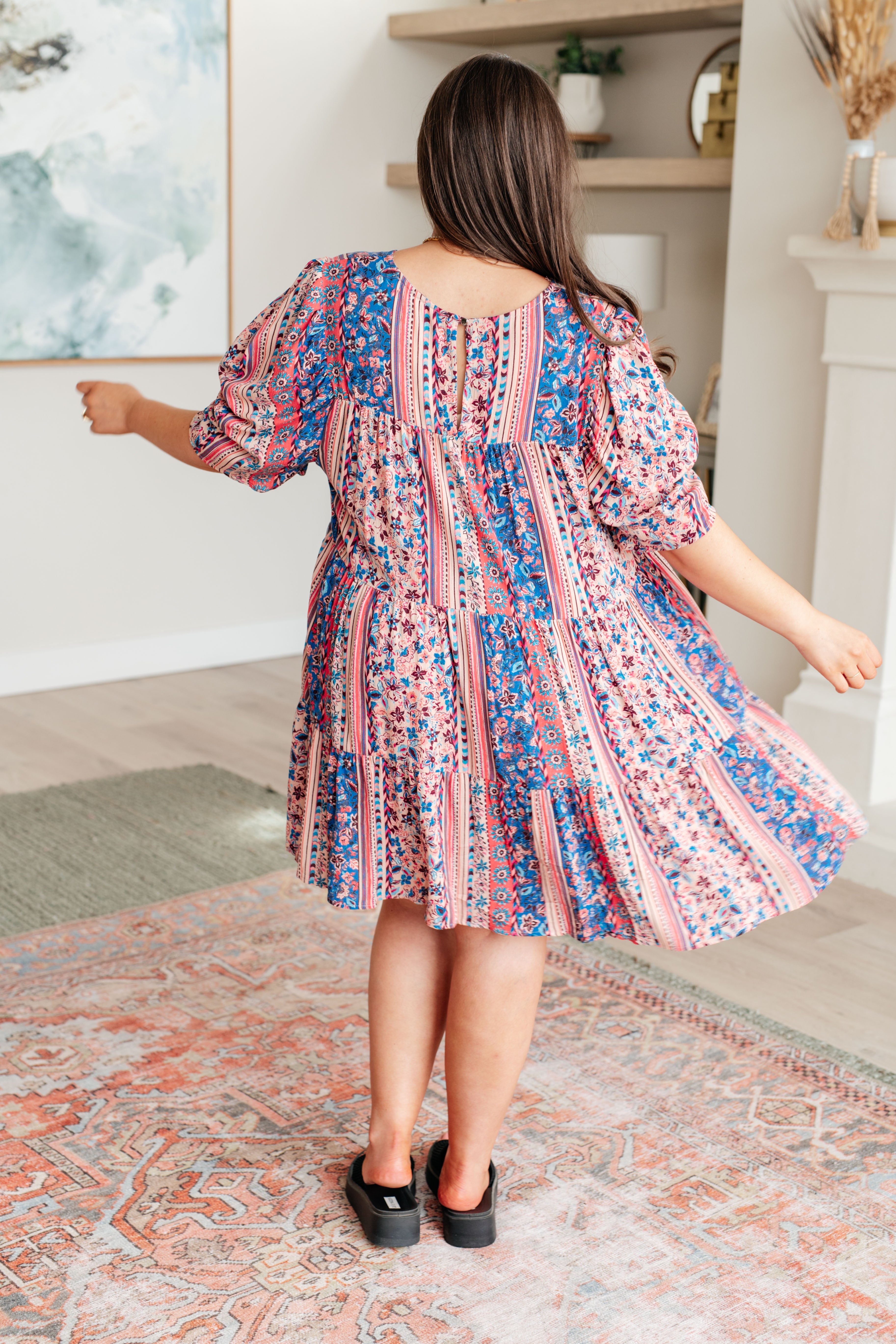 Rear view of woman wearing a pink and blue floral patterned dress with puffed sleeves and tiered skirt spinning indoors