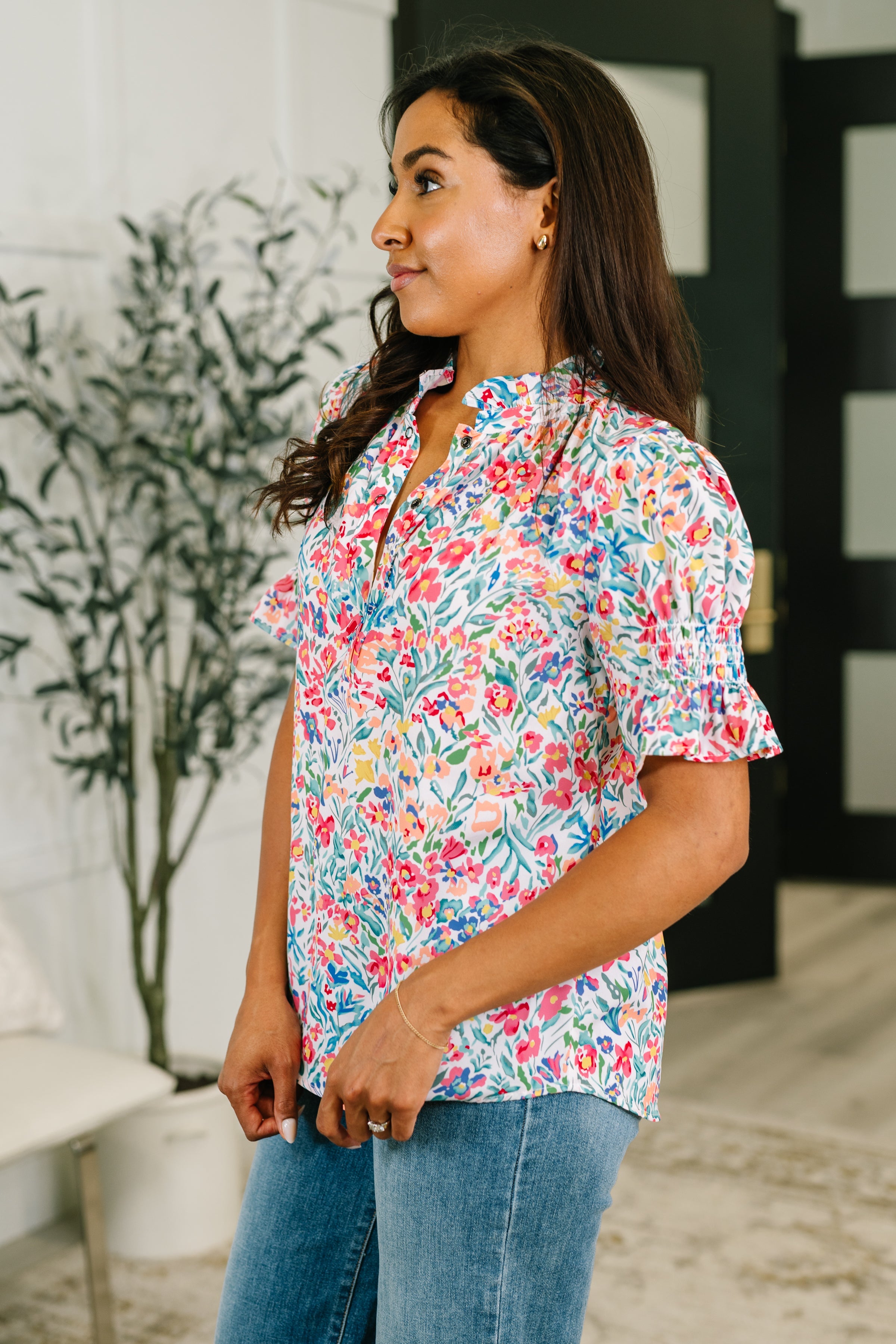 Woman wearing a floral split neck blouse in blue and coral paired with jeans in a bright, eclectic indoor setting