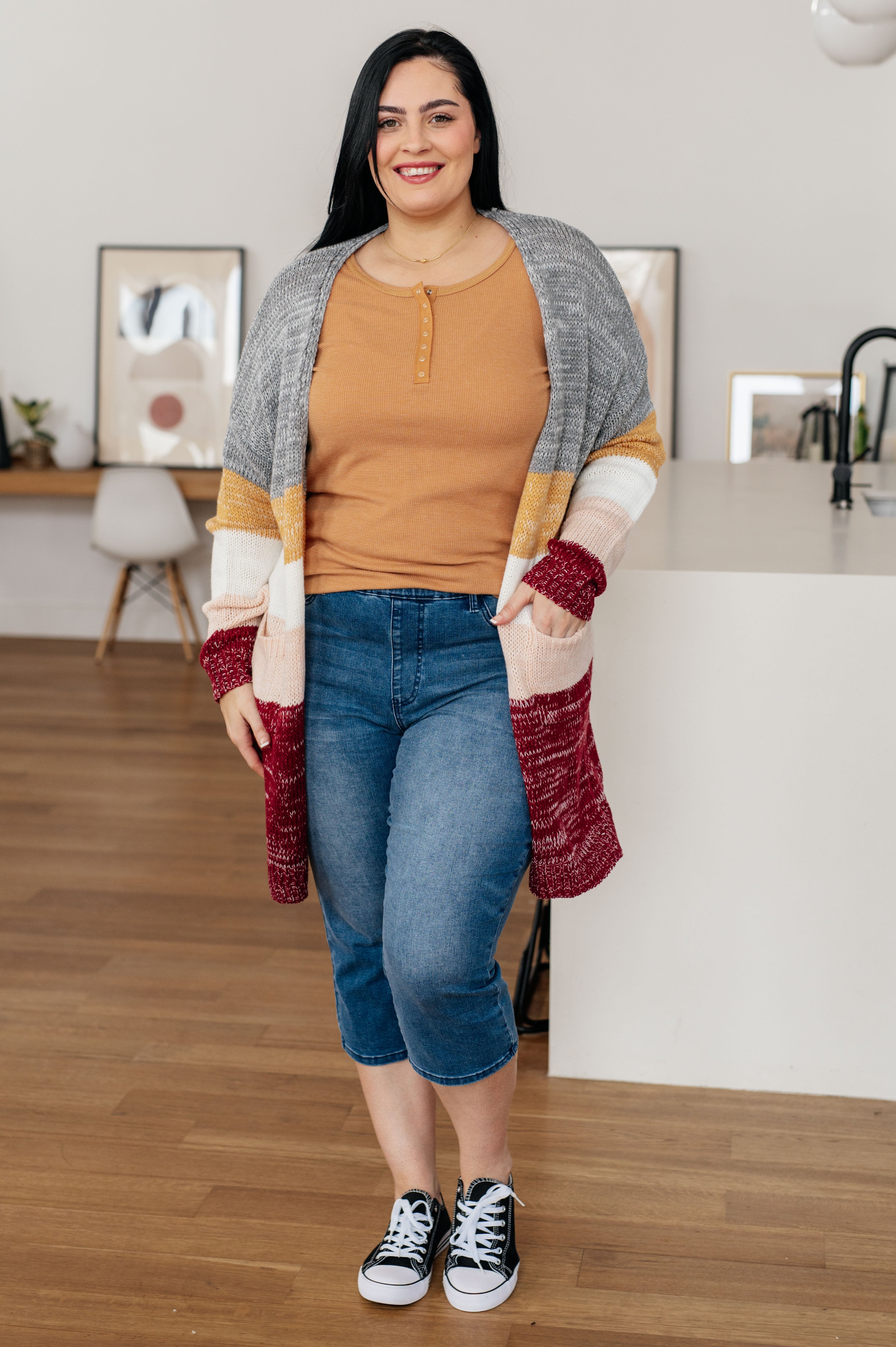 Smiling woman wearing a golden sand henley with snap buttons, a striped cardigan, cropped jeans, and black sneakers indoors