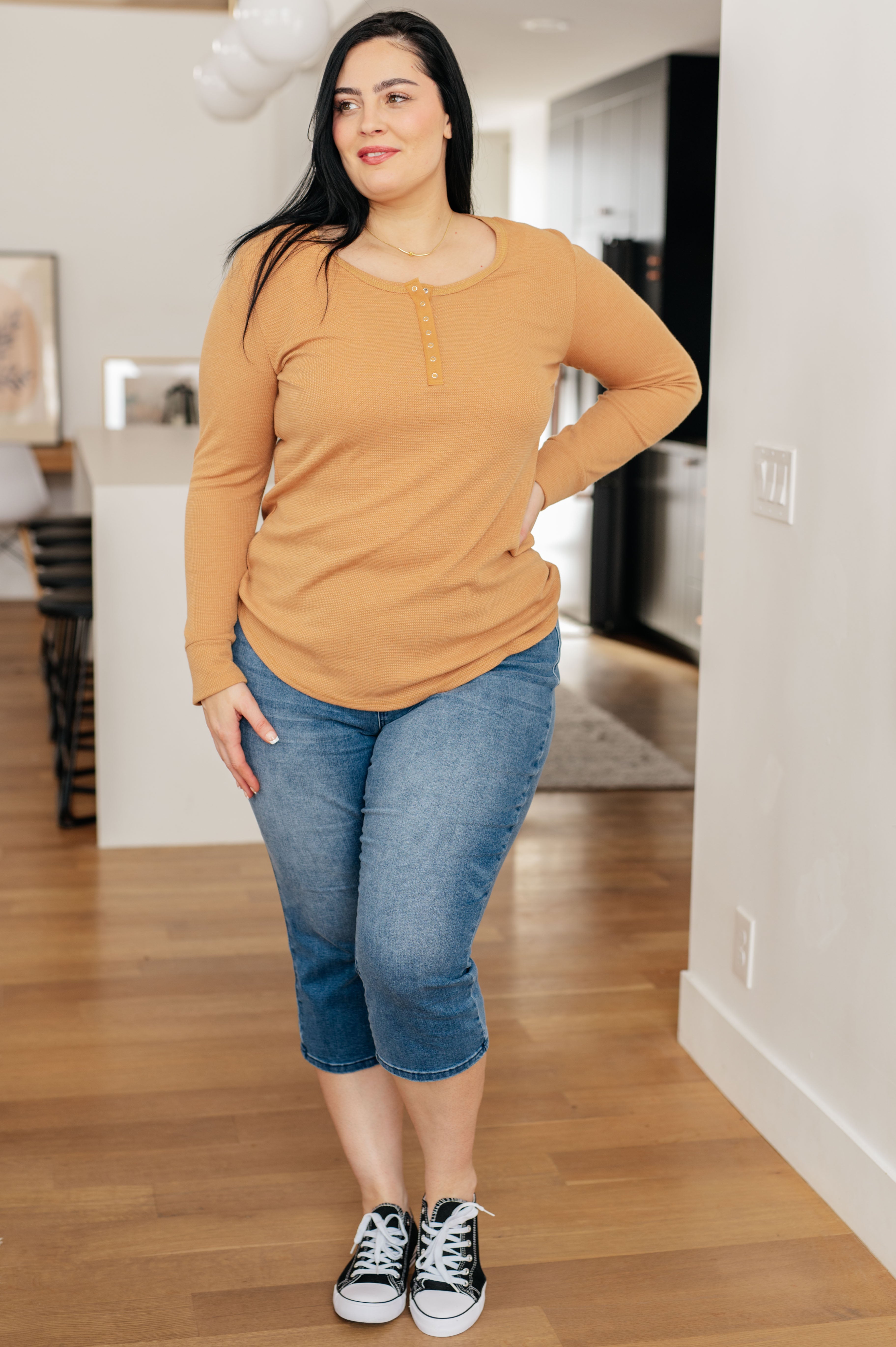 Woman with dark hair wearing a golden sand snap henley top and cropped blue jeans posing indoors on wood floor
