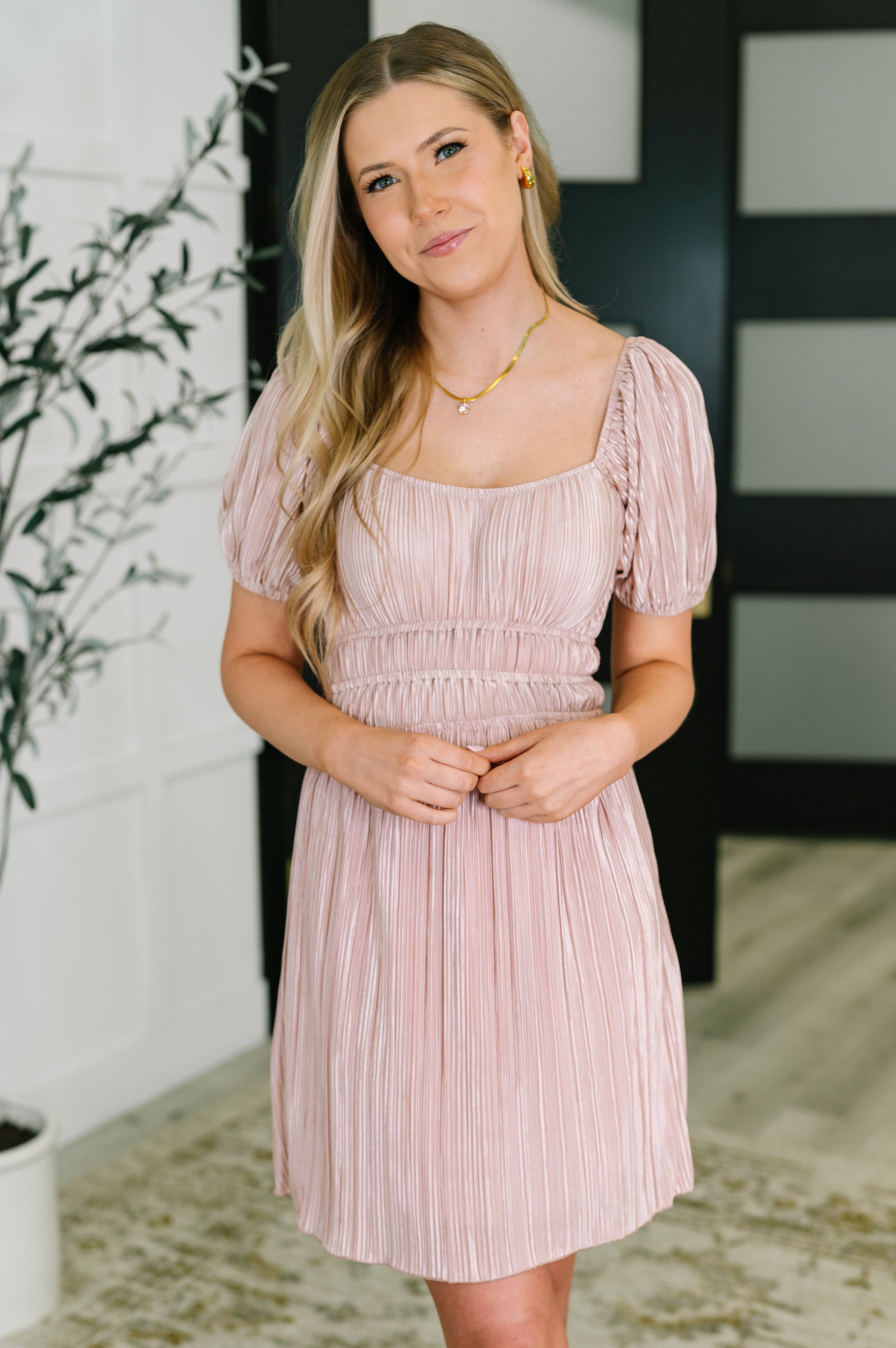 Woman wearing a pink pleated dress with puffed short sleeves and a square neckline standing indoors