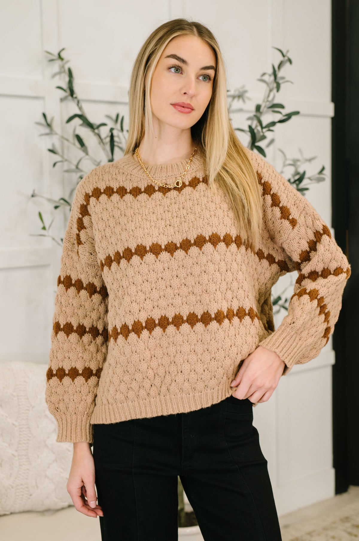 Woman modeling a textured brown sweater with patterned stripes and black pants in a bright indoor setting
