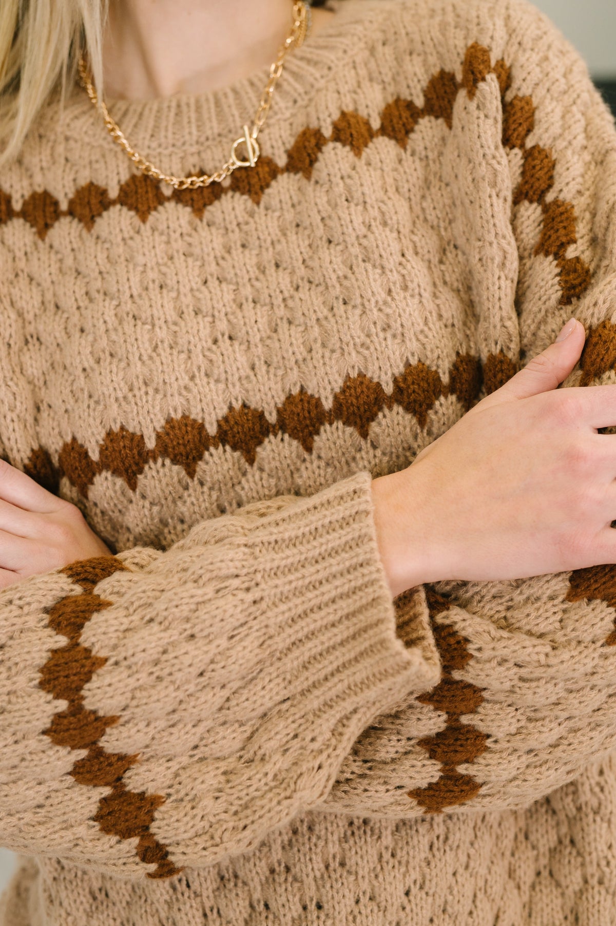 Close view of a woman wearing a taupe textured sweater with brown zigzag stripes and ribbed cuffs plus a gold chain necklace