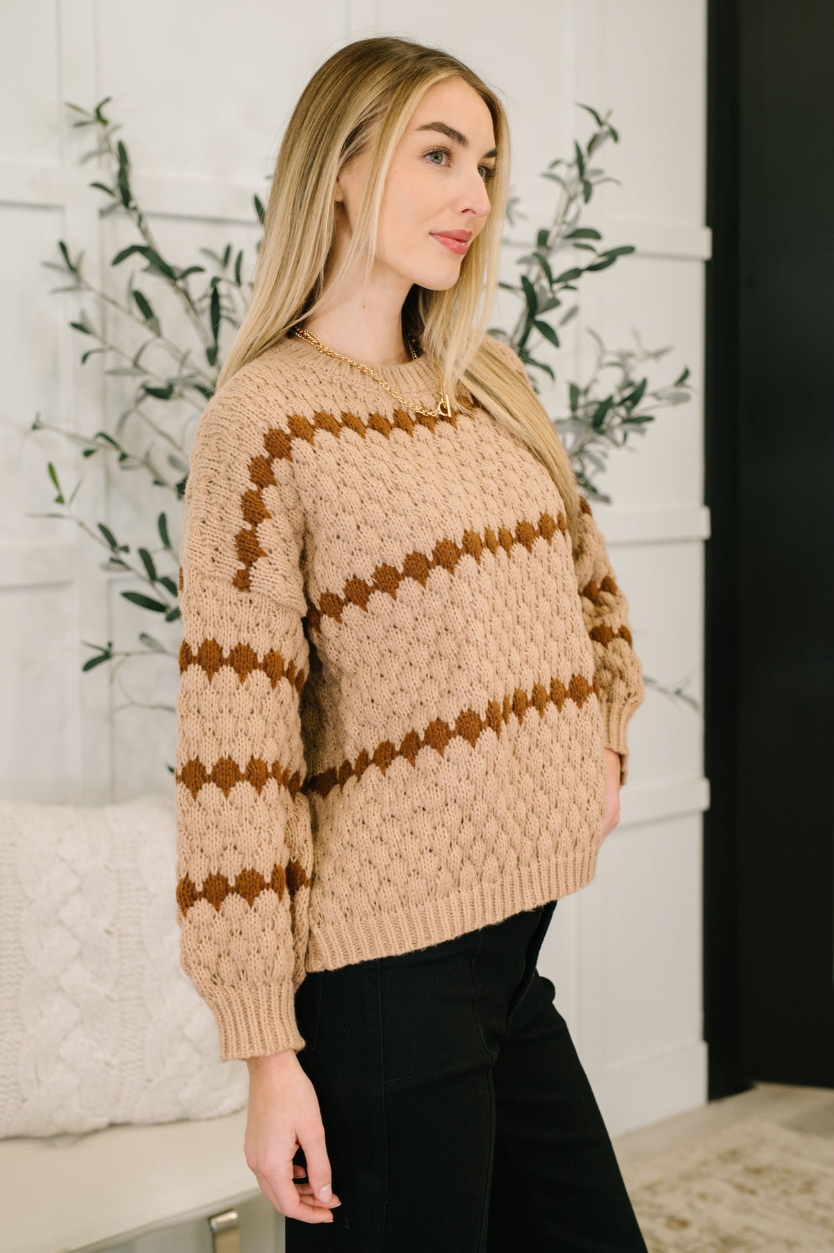 Woman wearing a taupe textured sweater with brown zigzag stripes, ribbed trim, black pants, and a delicate gold necklace indoors