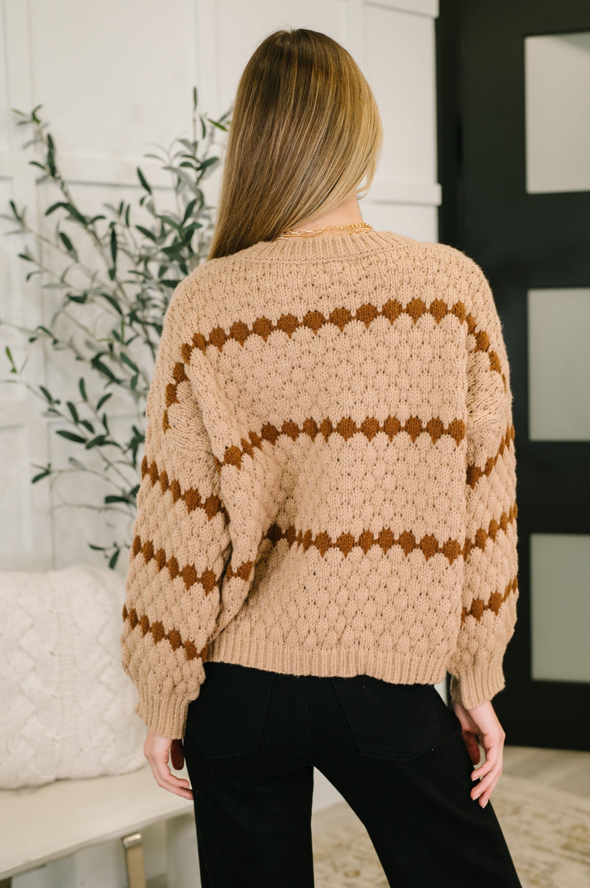 Back view of a woman wearing a taupe textured sweater with brown geometric stripes, ribbed cuffs and hem, and black jeans indoors