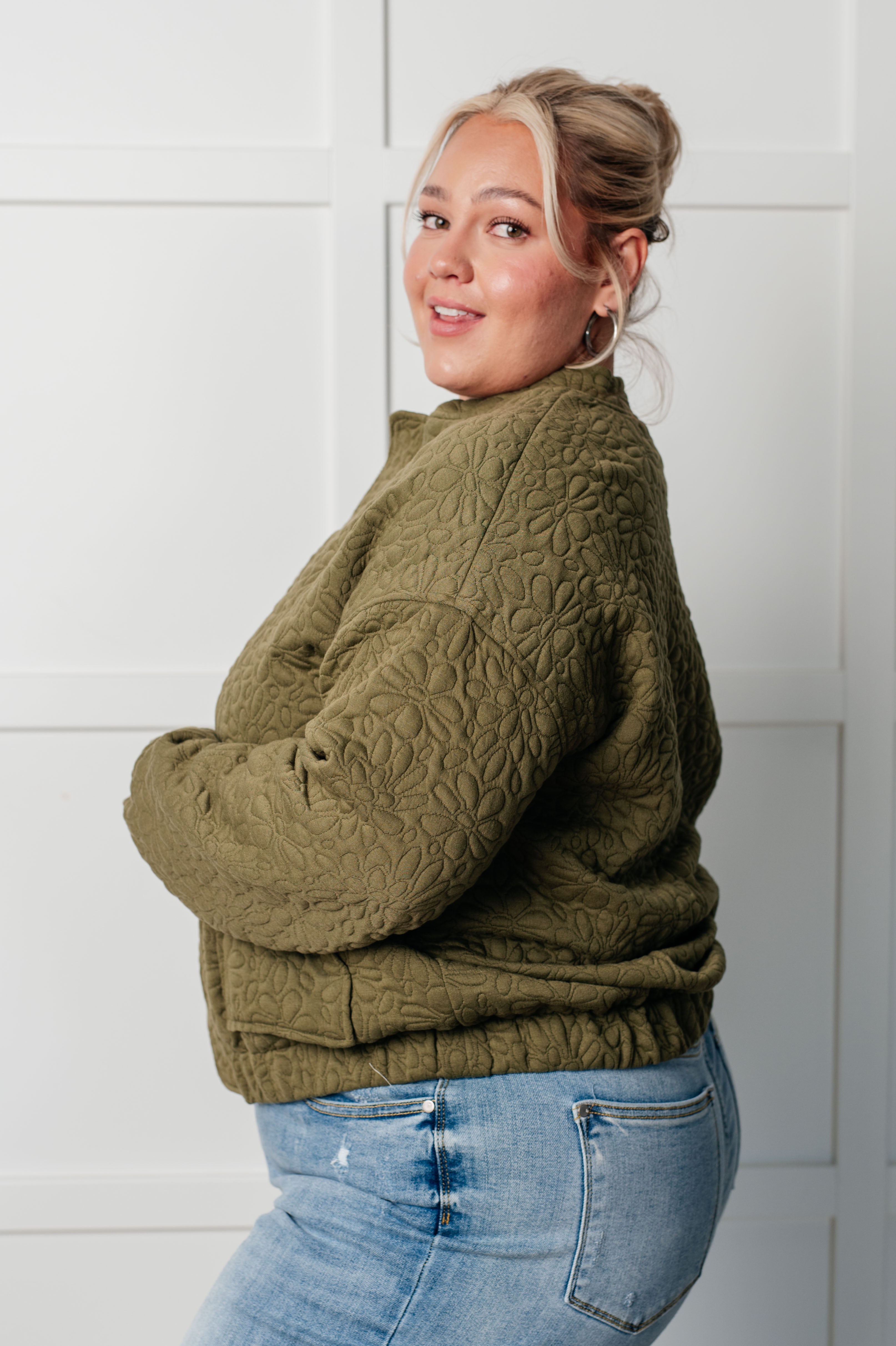 Woman wearing an olive green floral quilted zip up jacket and blue jeans standing against a white wall.