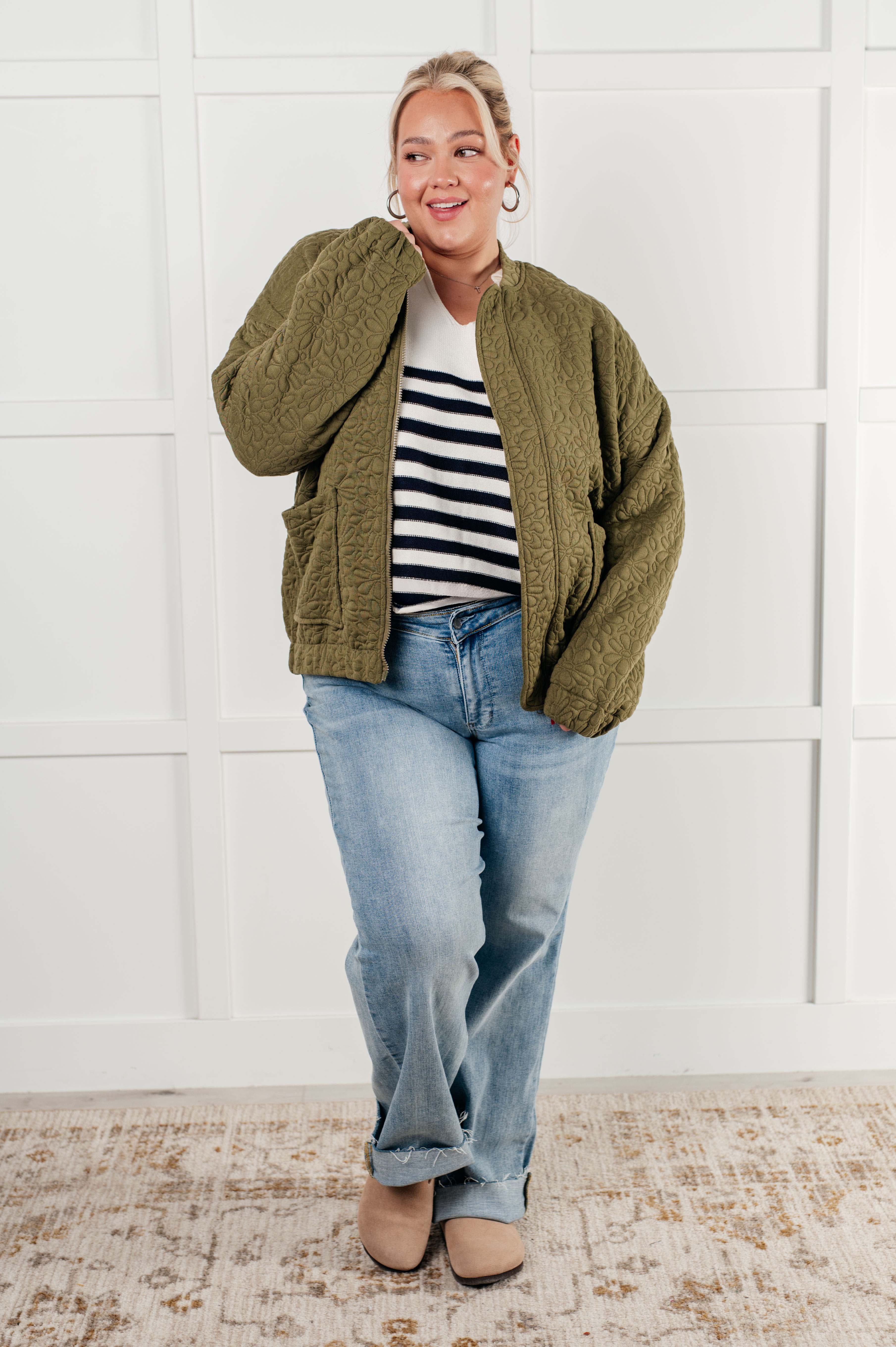 Woman wearing an olive green floral quilted zip up jacket over a black and white striped shirt, blue jeans and brown boots standing against a white wall.