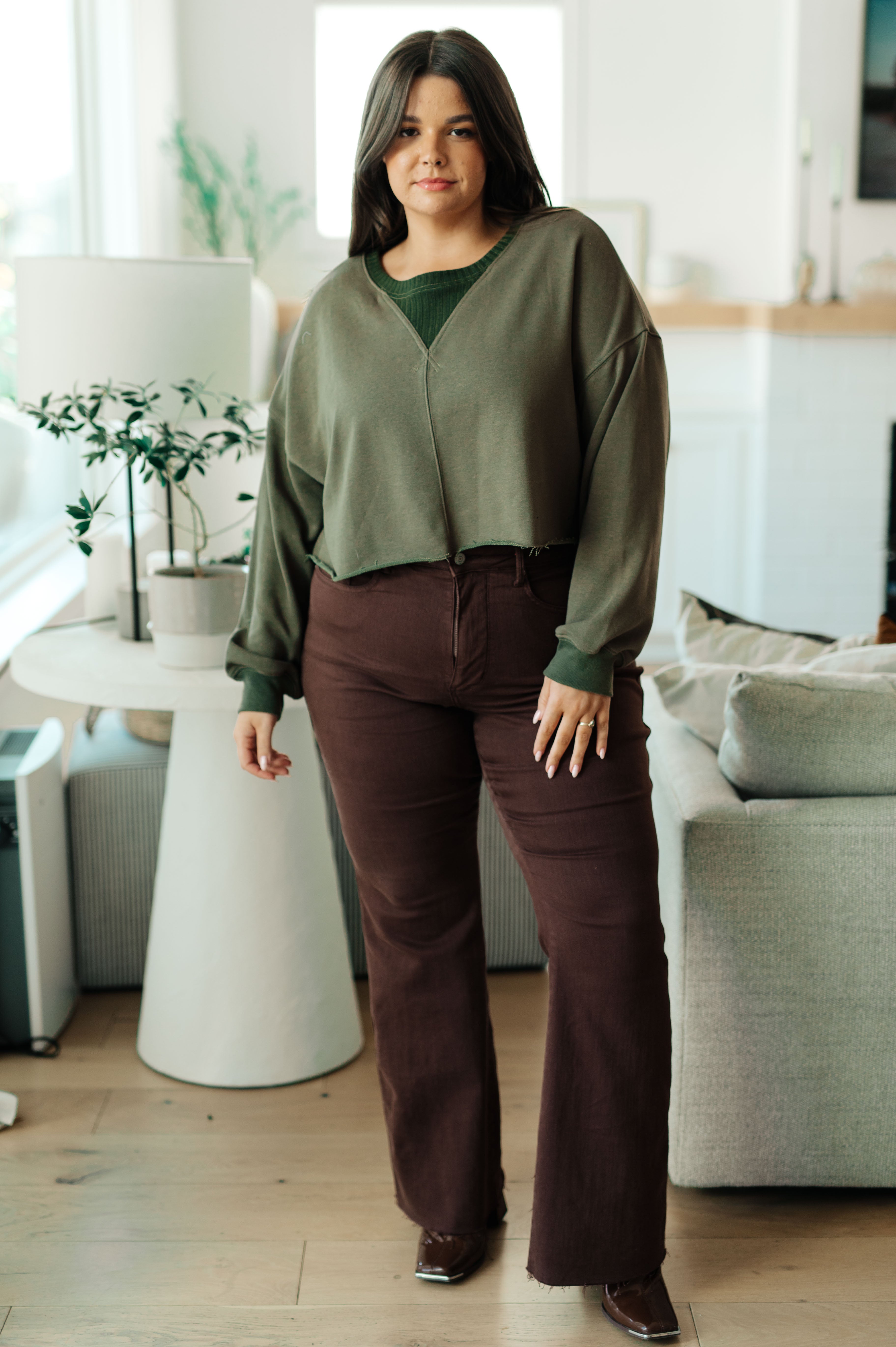 Woman indoors wearing green Just a Casual Crop Pullover layered over matching top paired with high-waisted brown pants