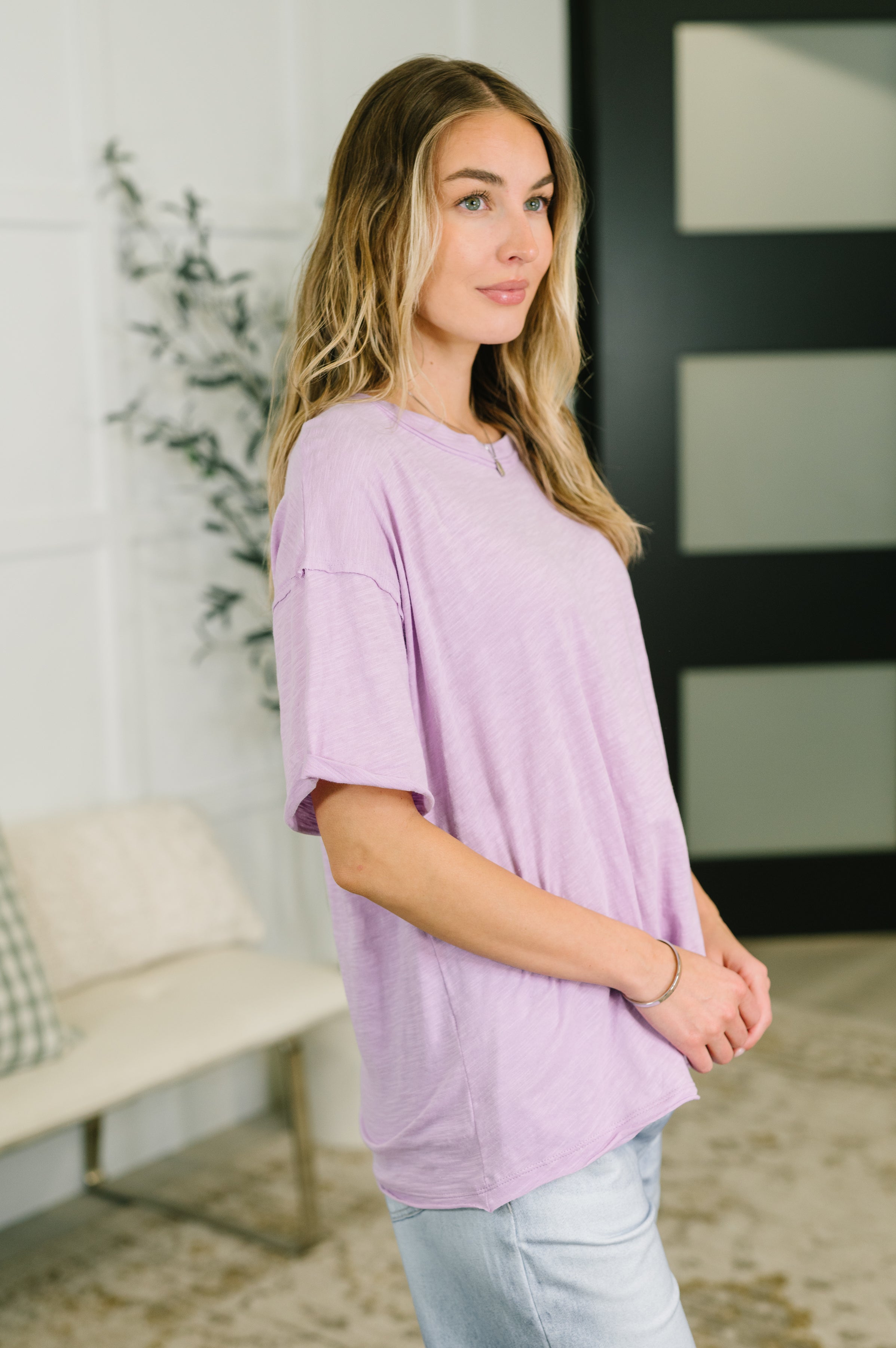 Woman wearing relaxed lilac tee with short sleeves and light wash jeans standing indoors near plant and door