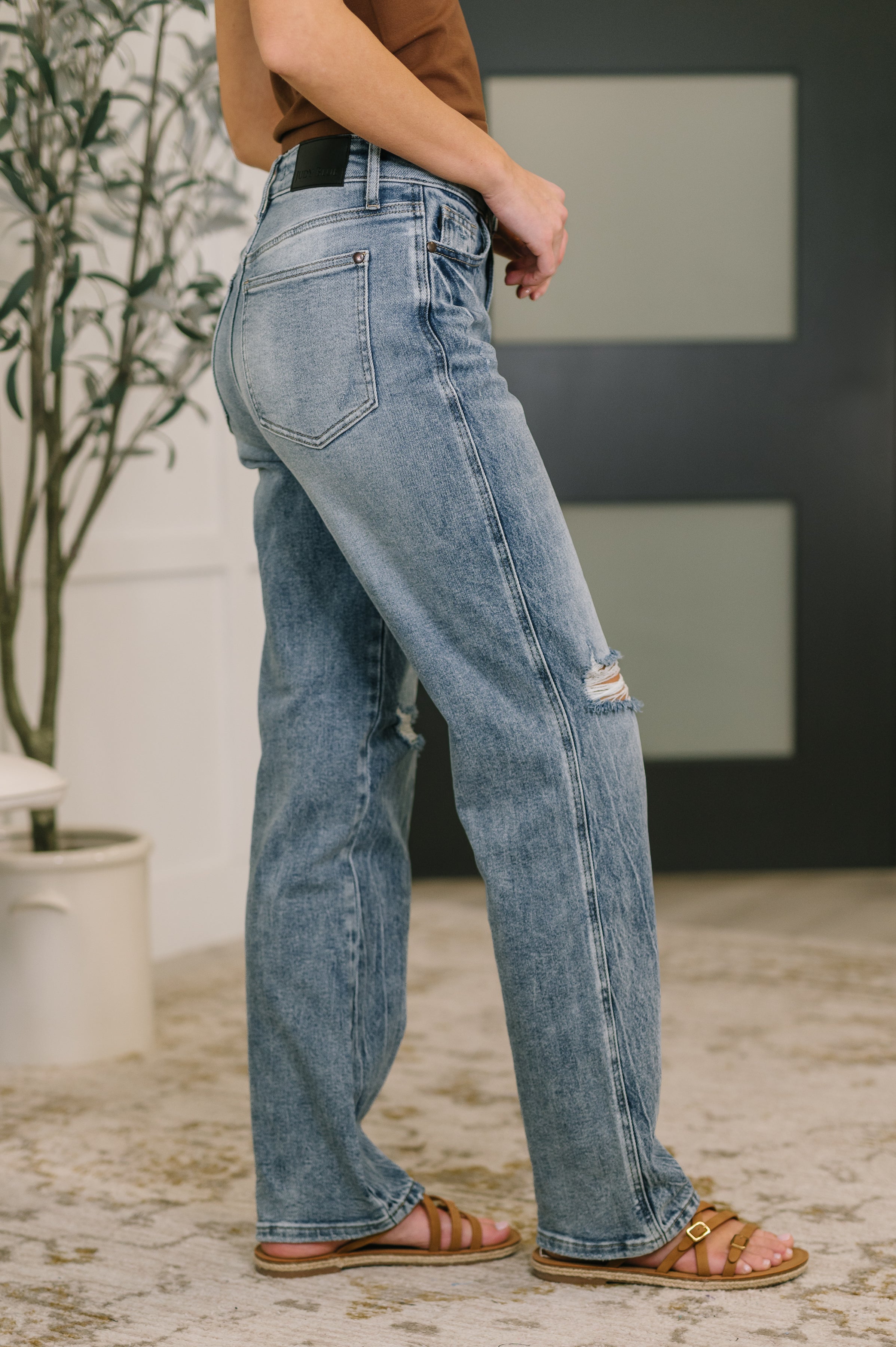 Side view of woman wearing high waist light blue rigid slim straight jeans with knee distressing and brown sandals indoors