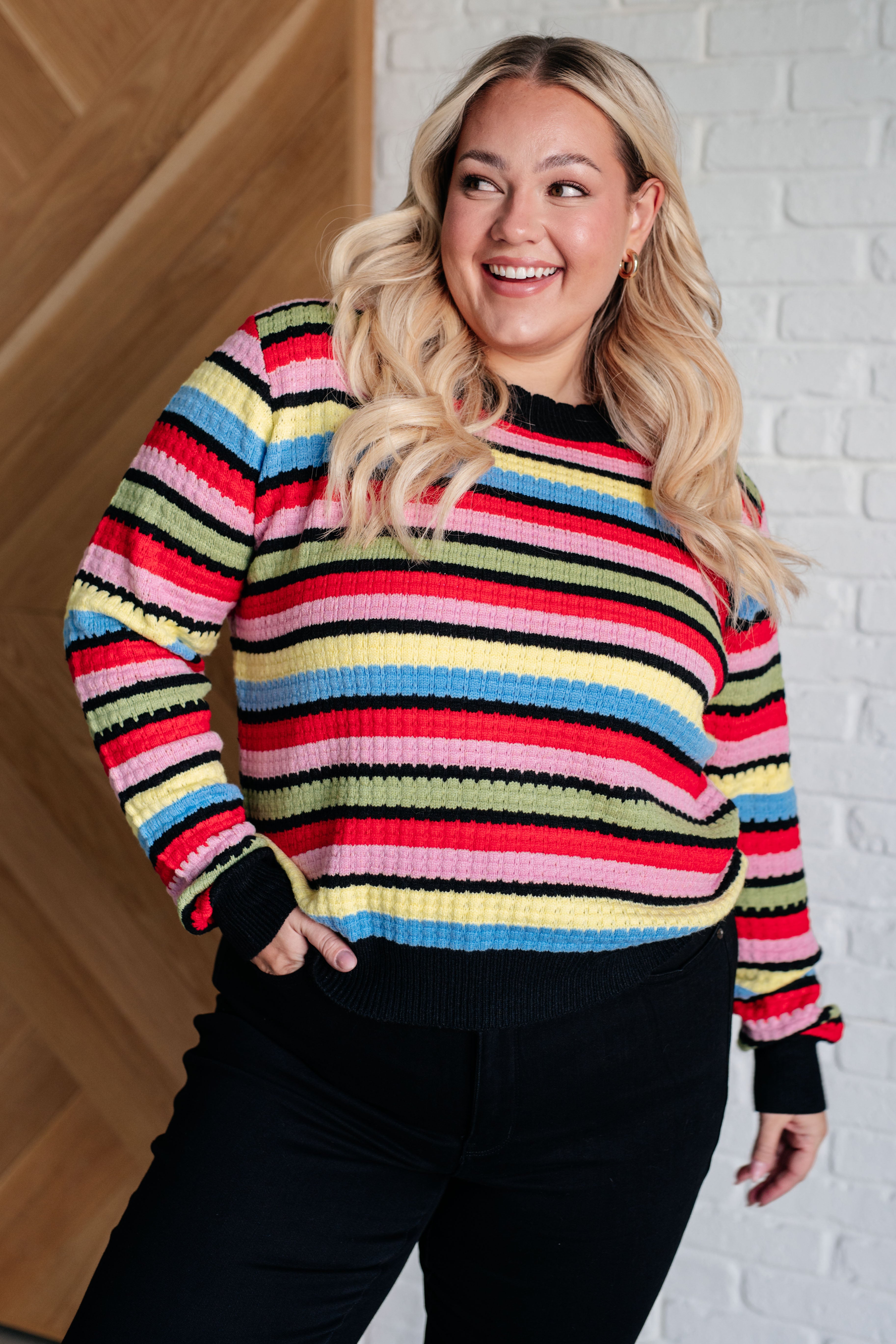Smiling woman indoors wearing multicolor Keep Dreaming Striped Sweater with black ribbed cuffs and hem paired with black pants
