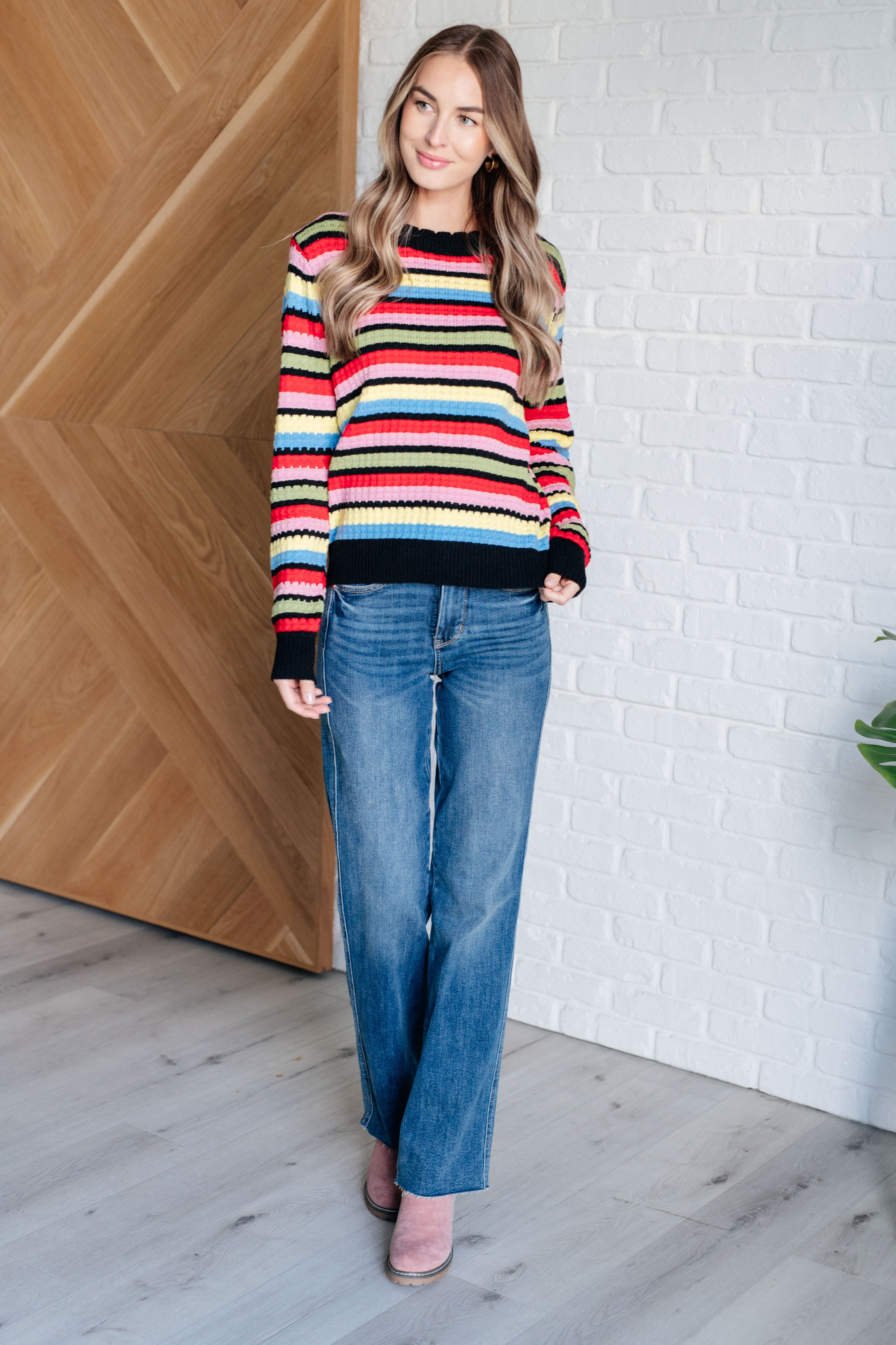 Woman standing indoors wearing multicolor Keep Dreaming Striped Sweater with black ribbed neckline and cuffs paired with blue jeans