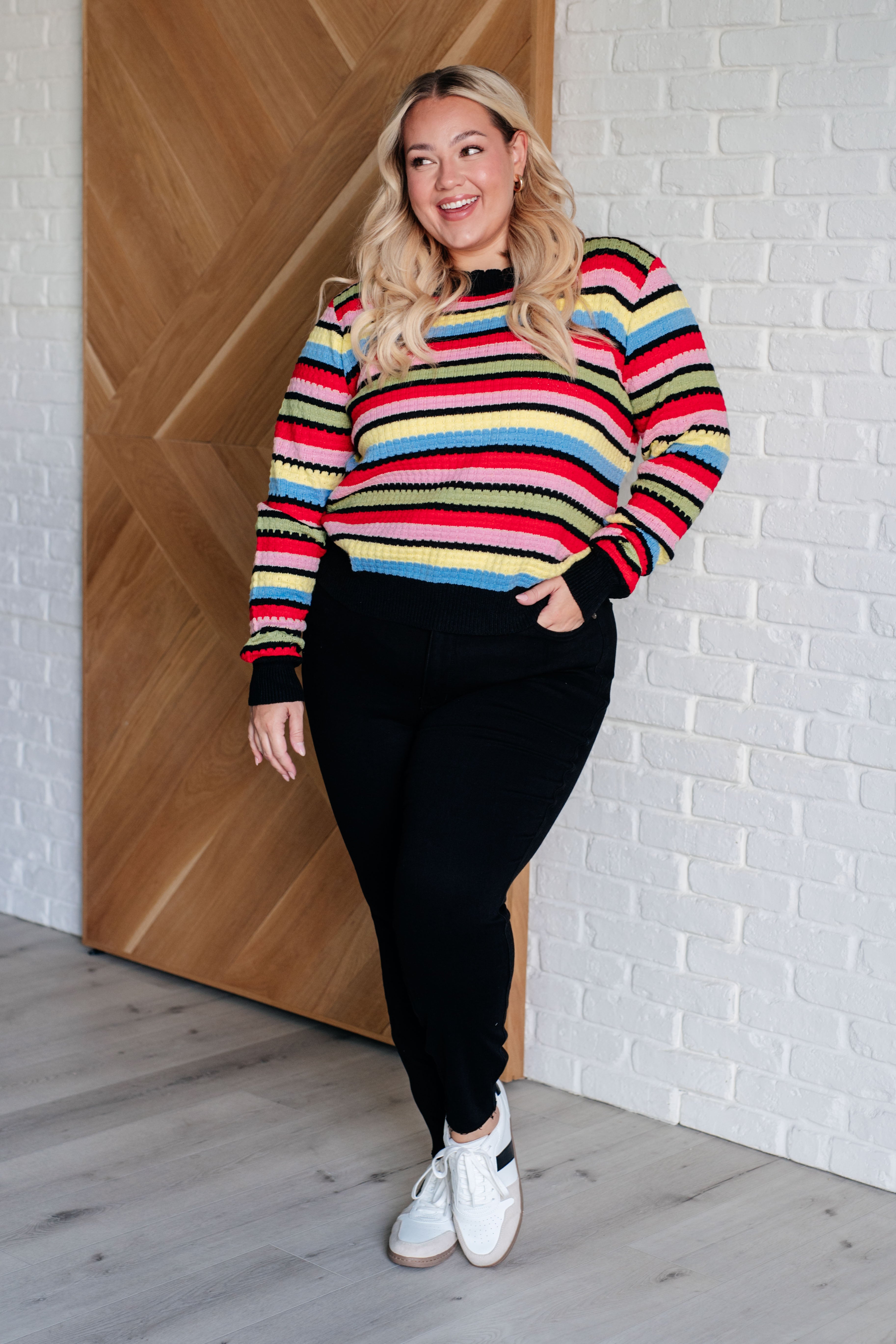 Woman standing indoors wearing multicolor Keep Dreaming Striped Sweater with black cuffs and hem paired with black pants and white sneakers