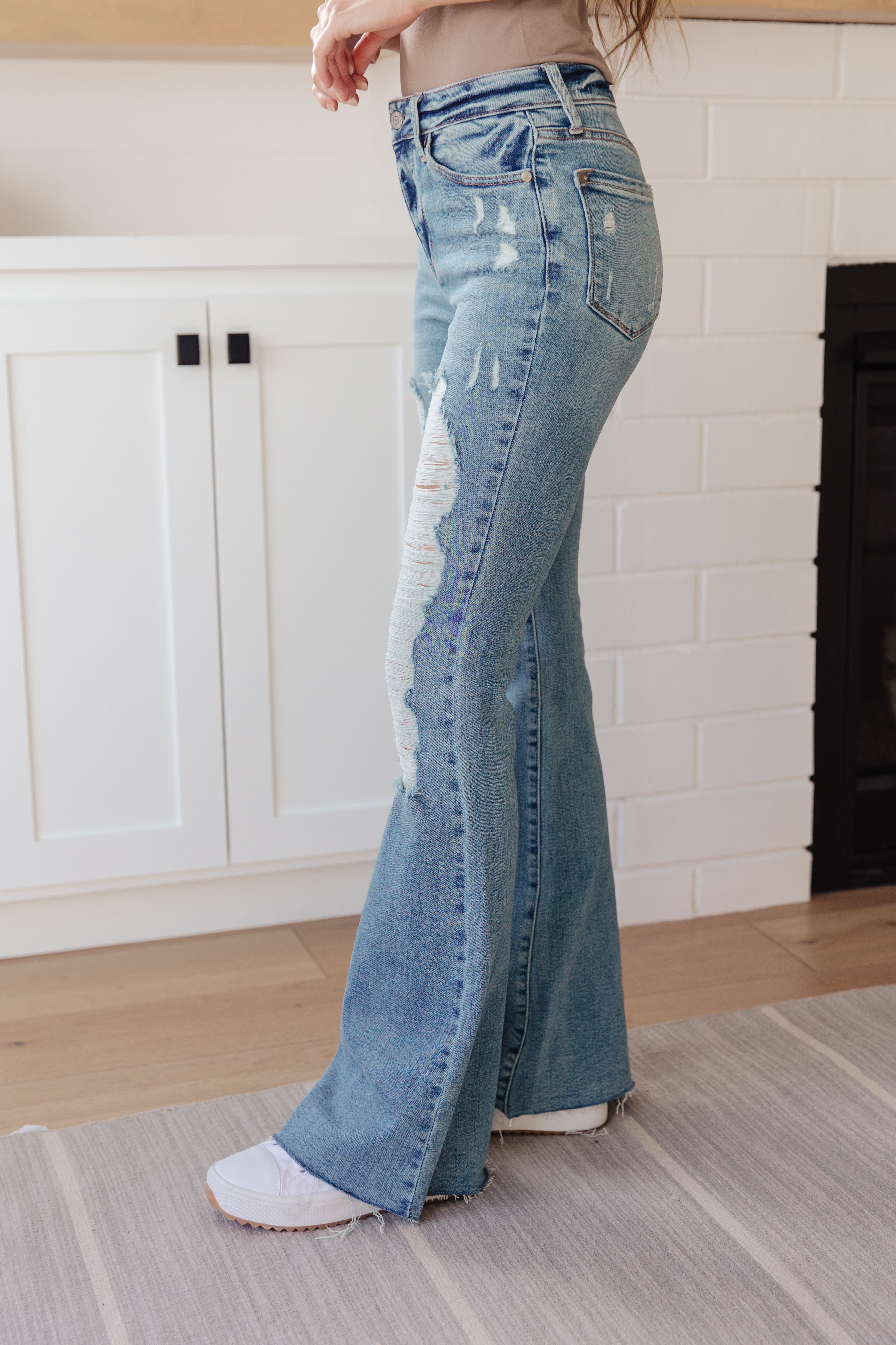 Side view of person wearing light blue high-rise flare jeans with heavy distressing and white sneakers indoors on a rug