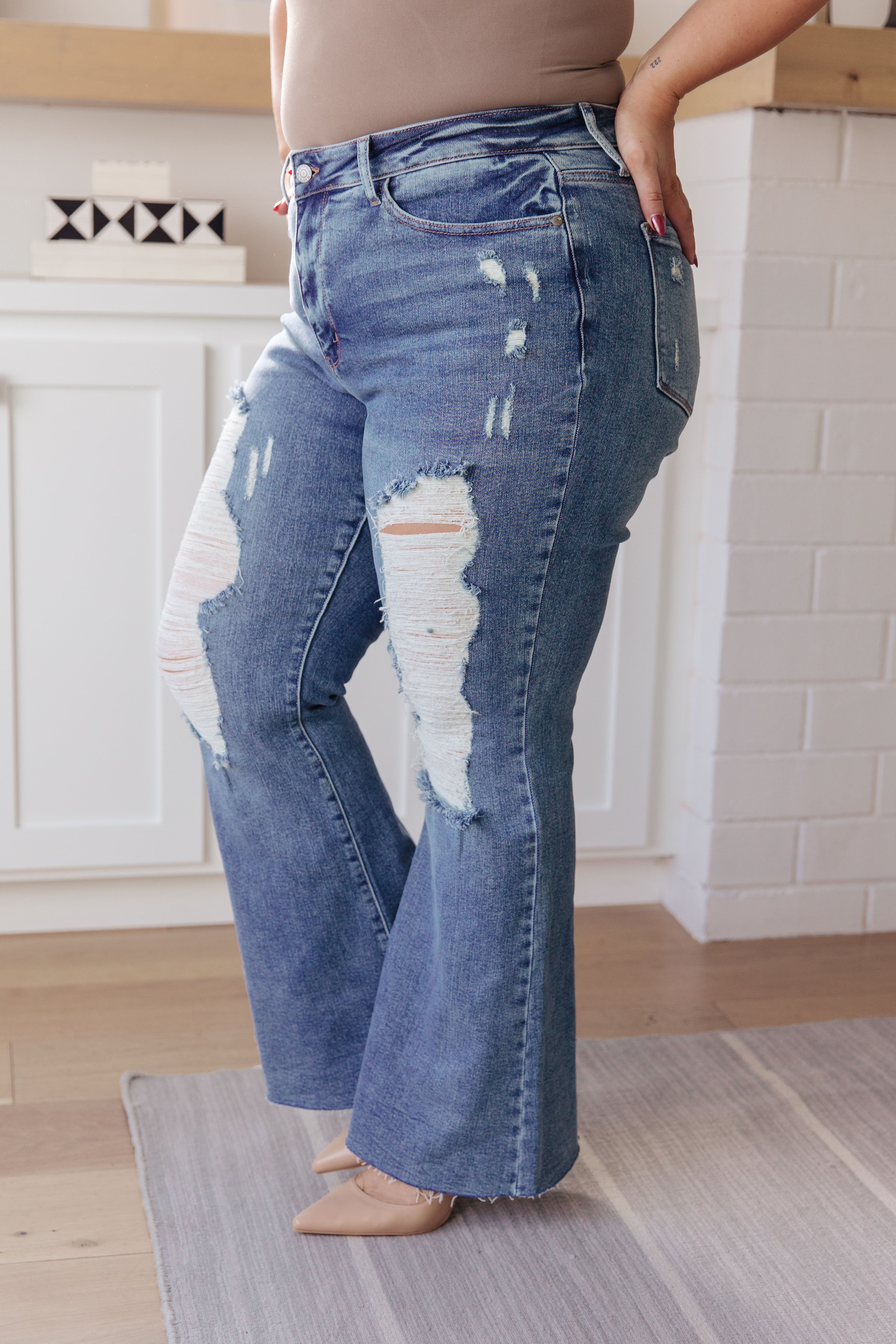 Plus-size person wearing blue high-rise flare jeans with heavy distressing and nude pointed-toe heels indoors