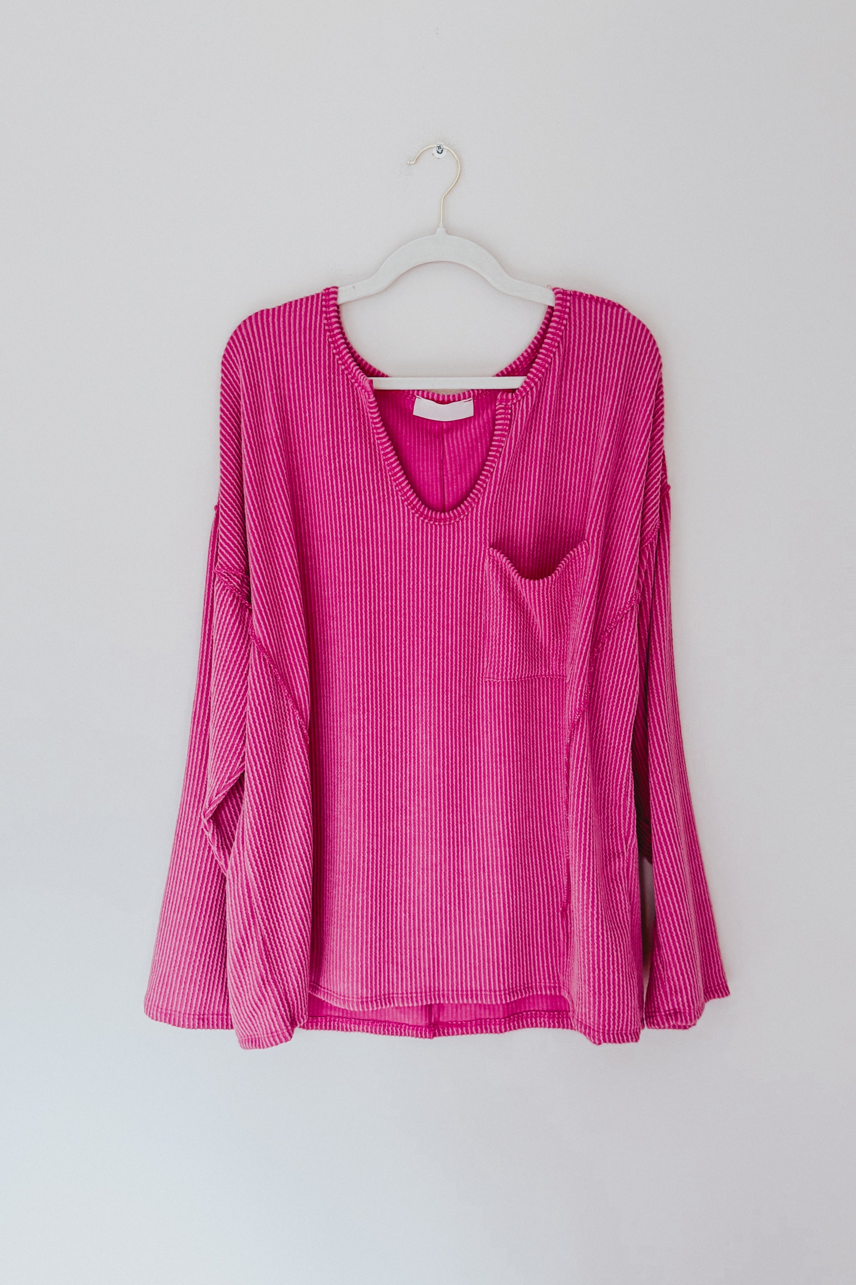Magenta ribbed long sleeve top with loose fit, scoop neckline, and front pocket hanging on white hanger against plain wall