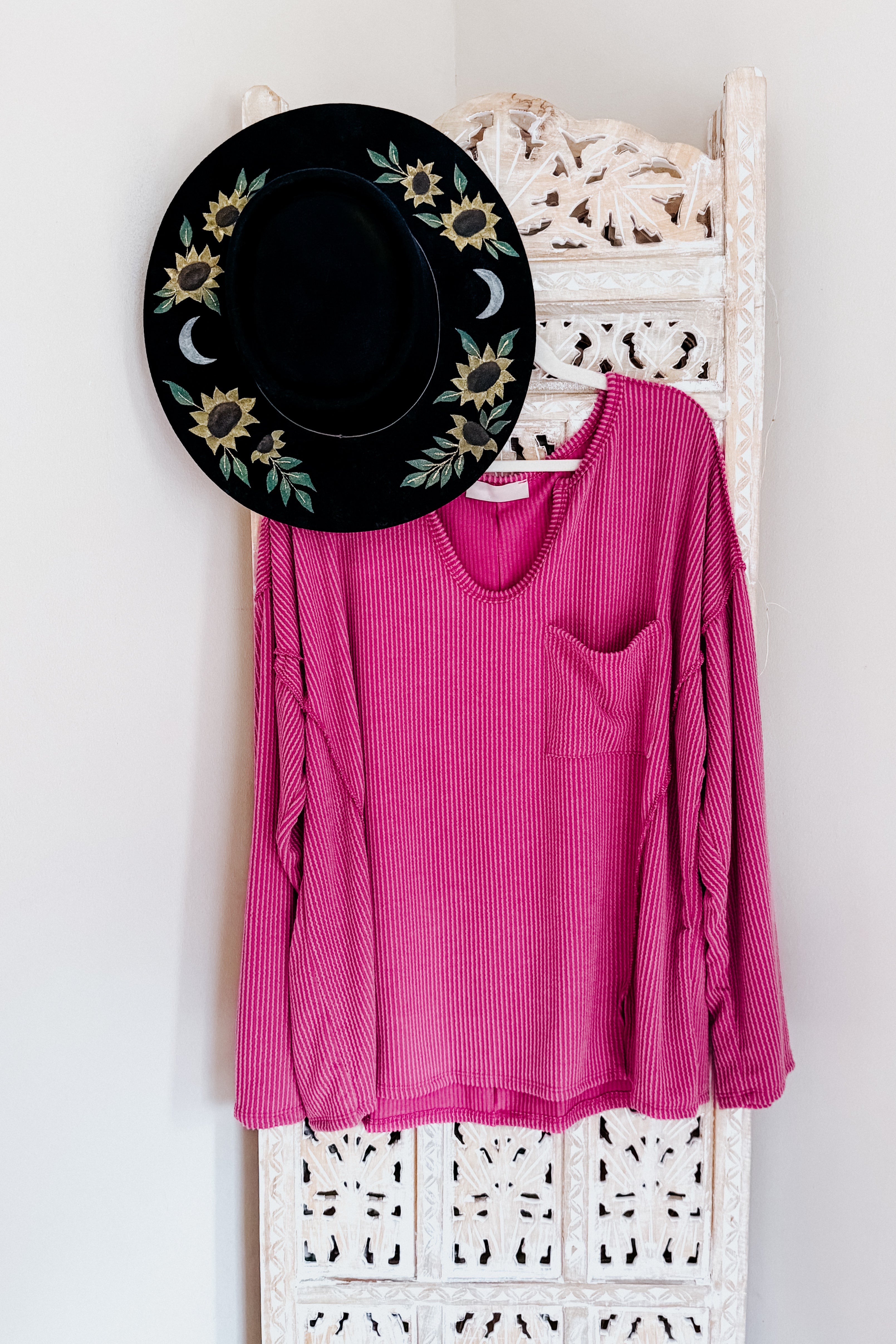 Magenta ribbed long sleeve top with front pocket hanging on carved white wooden screen with black floral hat above