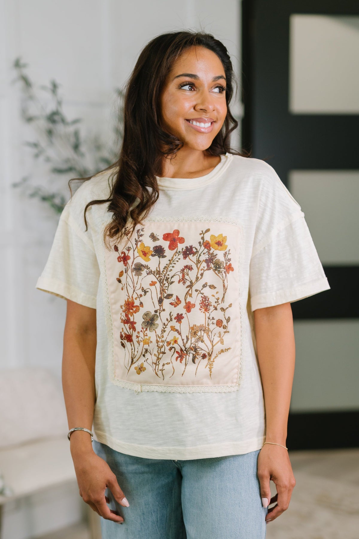 Smiling woman wearing a relaxed cream knit top with colorful floral applique panel and light wash jeans indoors