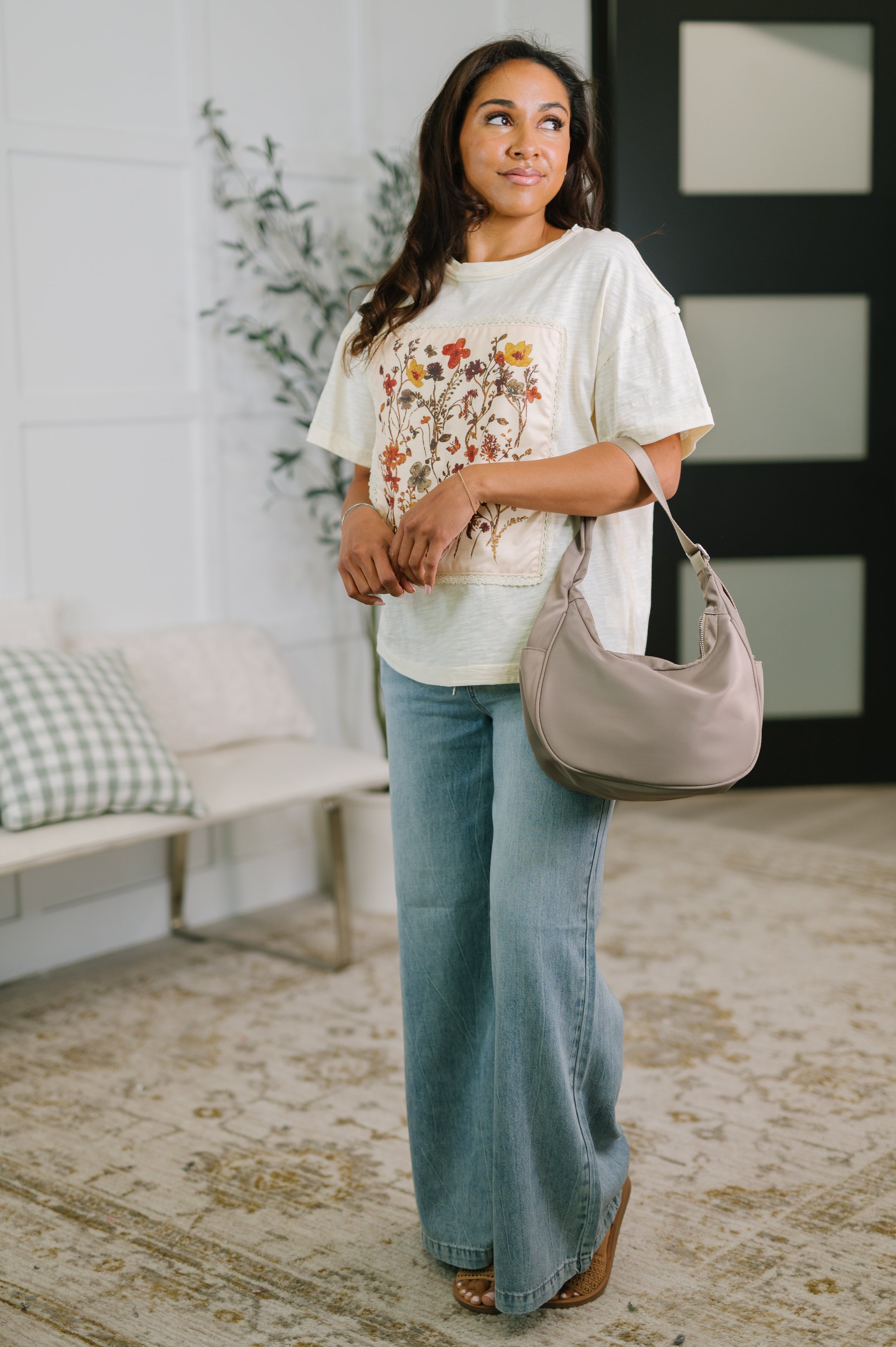 Woman wearing a cream floral applique top with relaxed short sleeves wide leg jeans and a taupe shoulder bag indoors