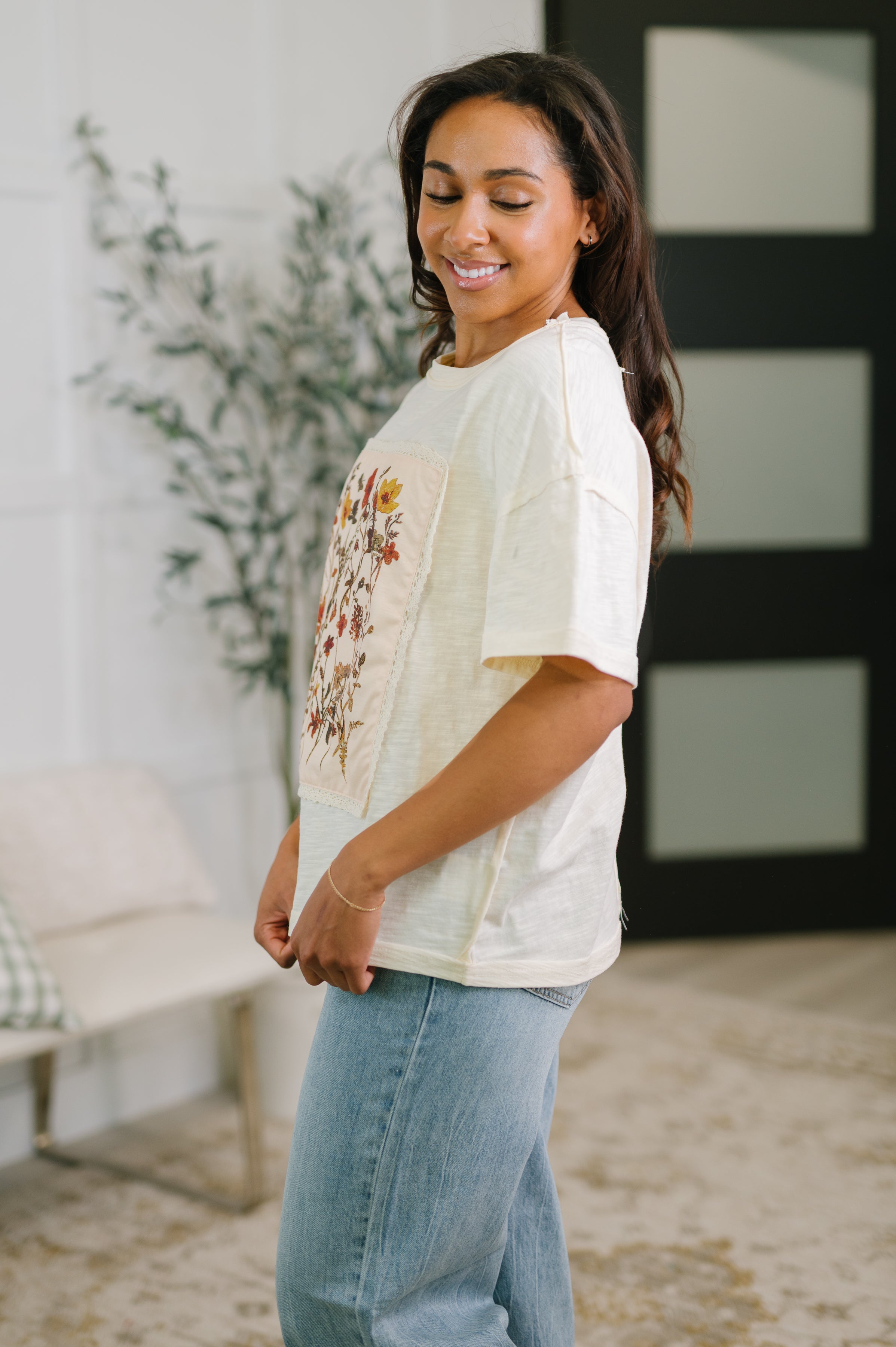 Woman smiling in profile wearing a cream floral applique top with short sleeves and light wash jeans indoors