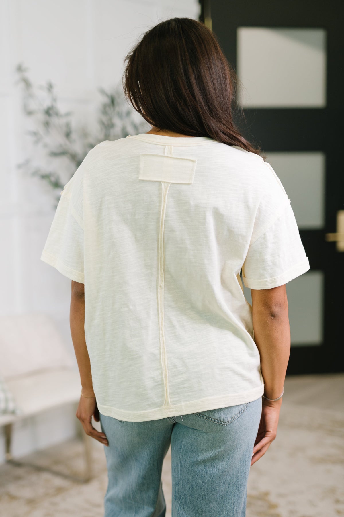 Back view of a woman wearing a cream short sleeve knit top with a center seam detail and light wash jeans indoors