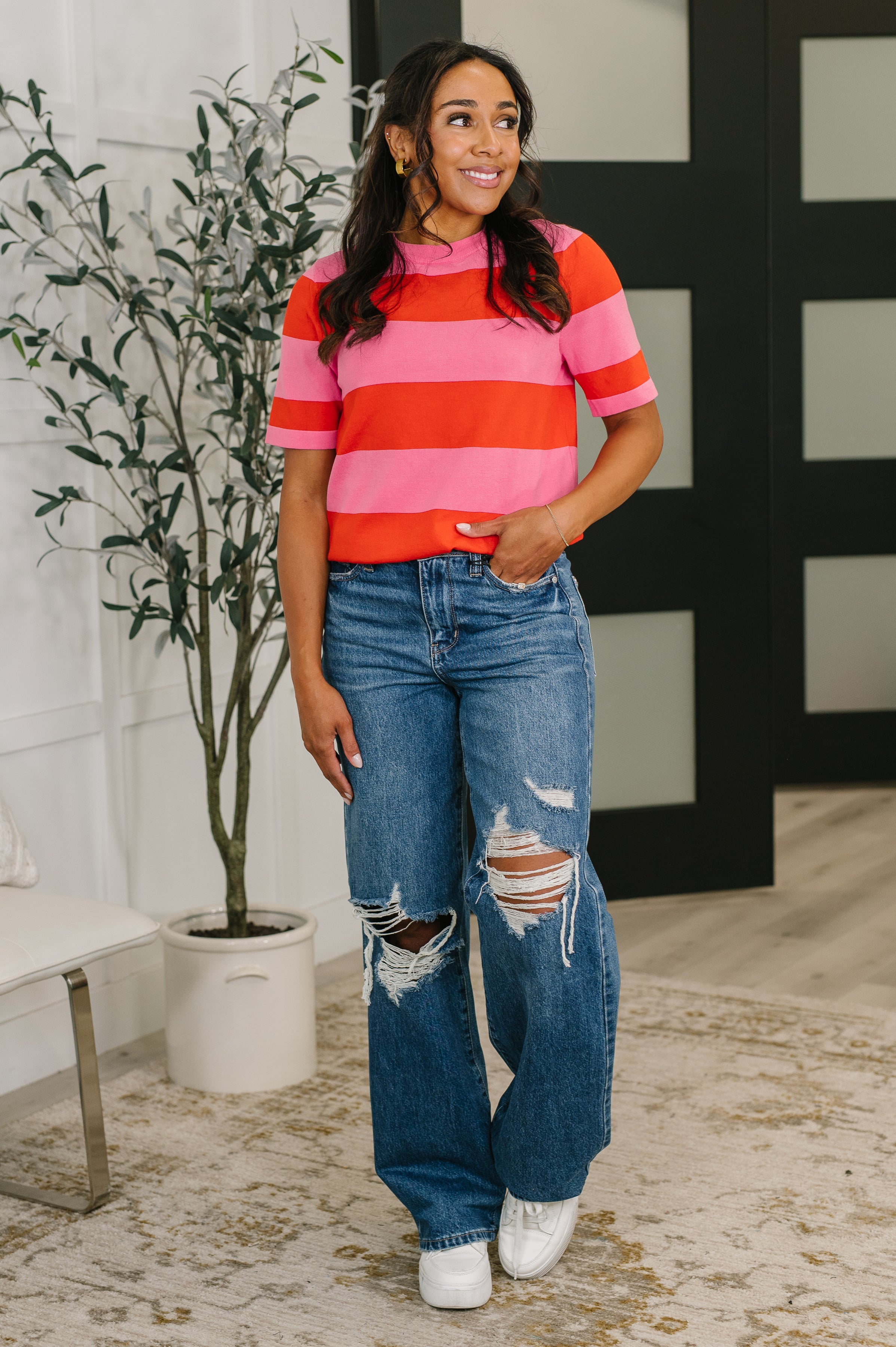 Woman wearing red and pink striped knit blouse paired with ripped wide-leg jeans and white sneakers indoors