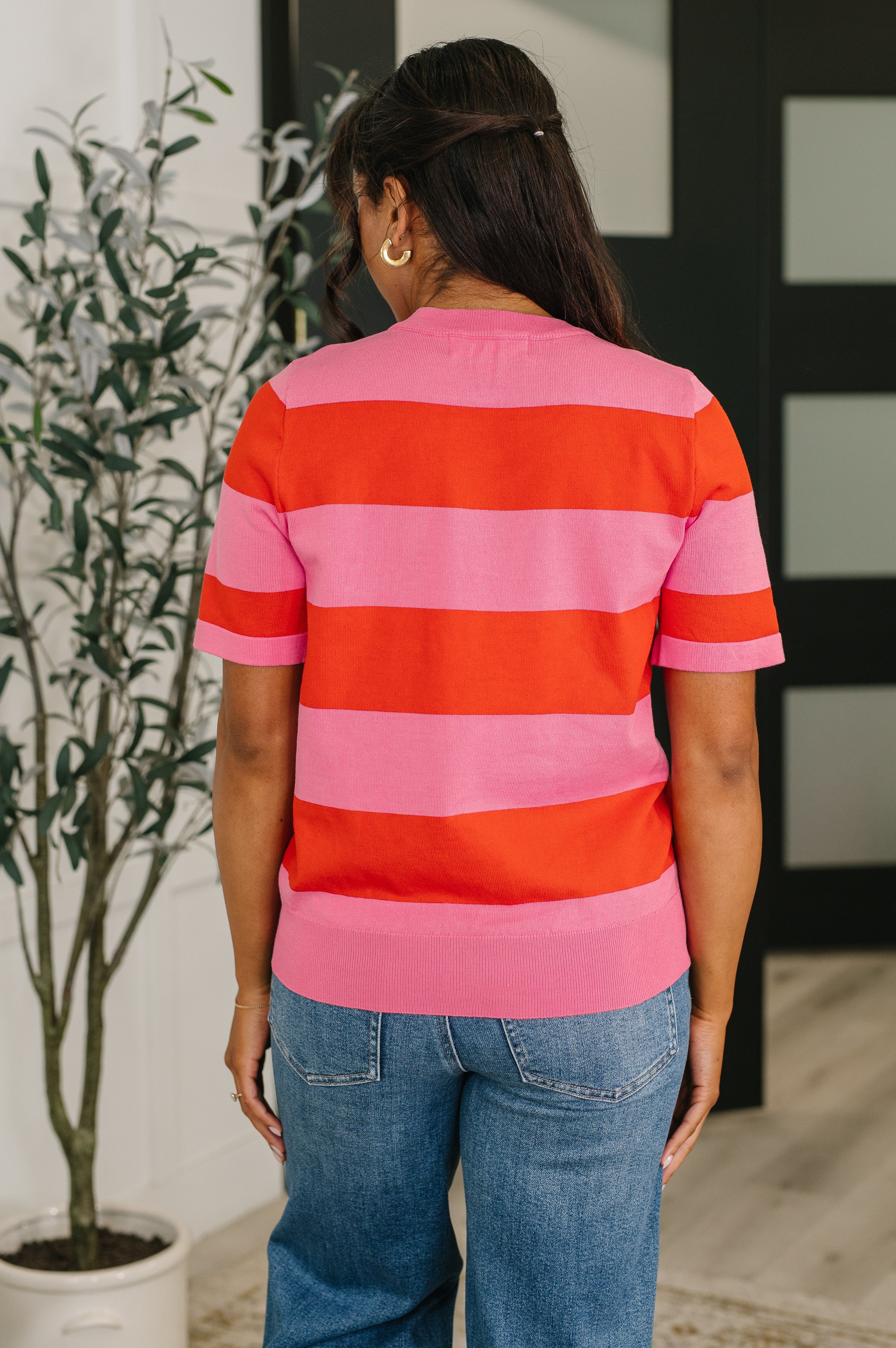 Rear view of woman wearing a pink and red striped knit blouse with short sleeves and relaxed blue jeans indoors