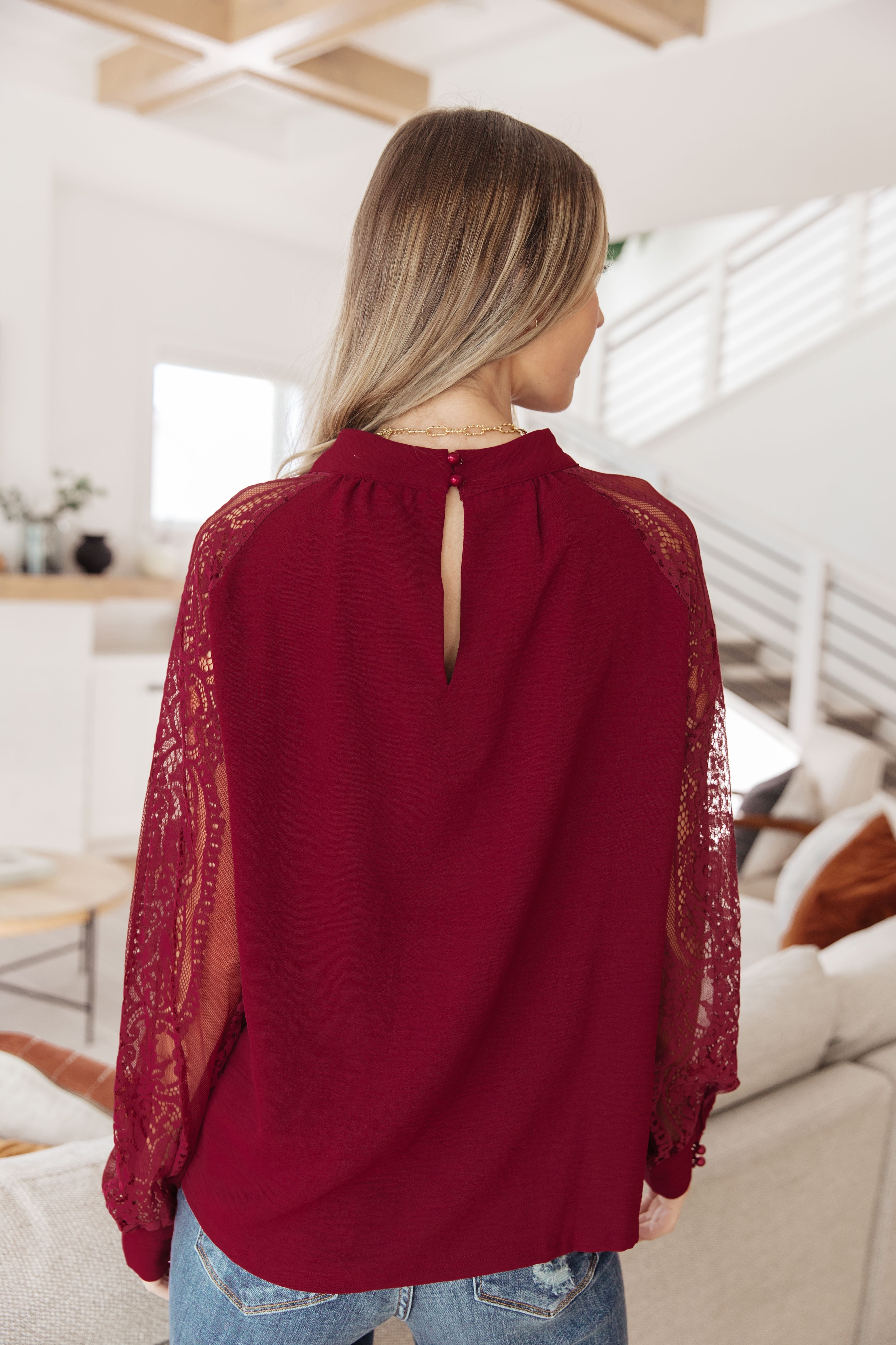 Back view of a woman wearing a wine blouse with lace bishop sleeves and a small button keyhole closure at the neckline indoors