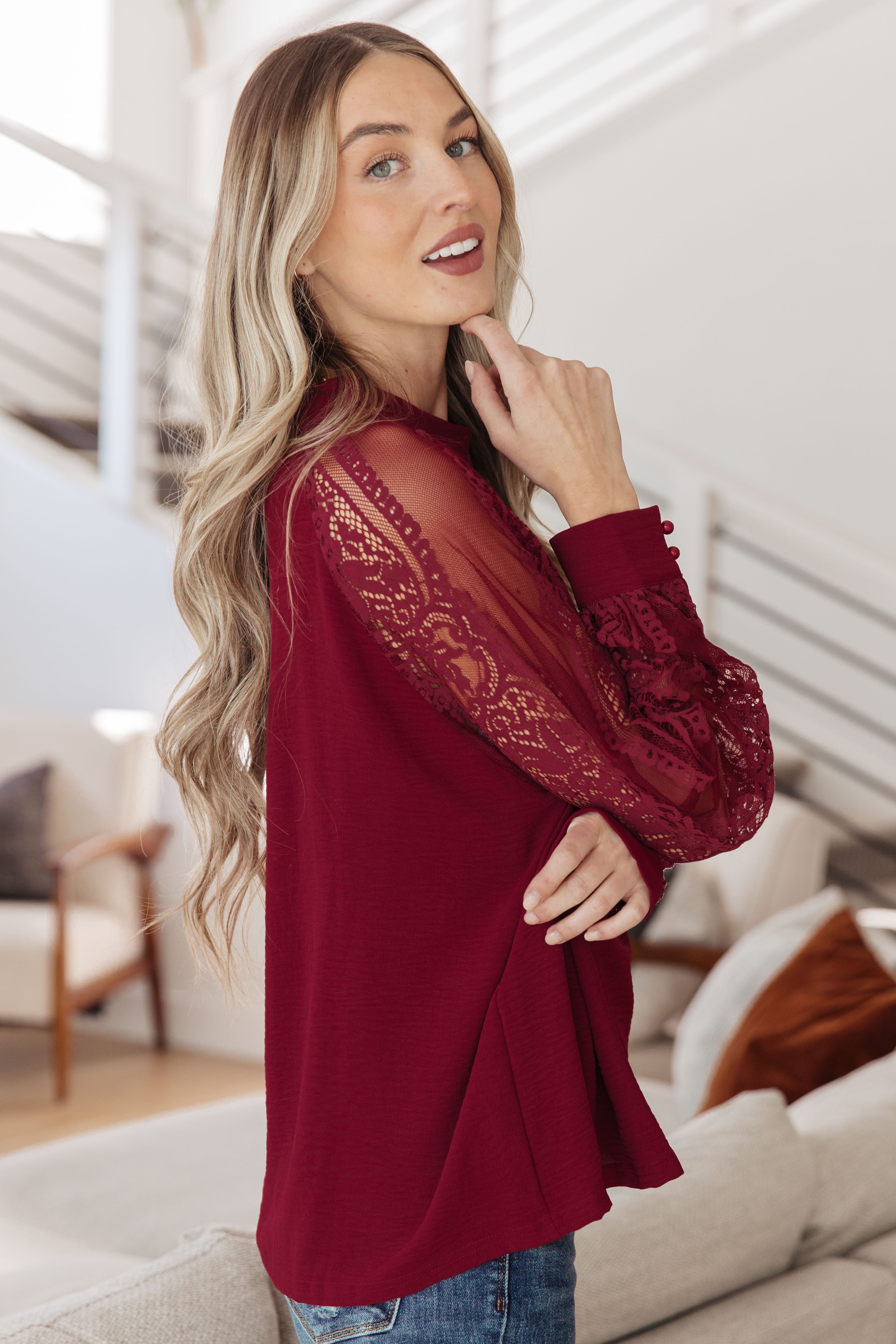 Woman wearing a wine-colored blouse with intricate lace sleeves and buttoned cuffs posing indoors in casual jeans