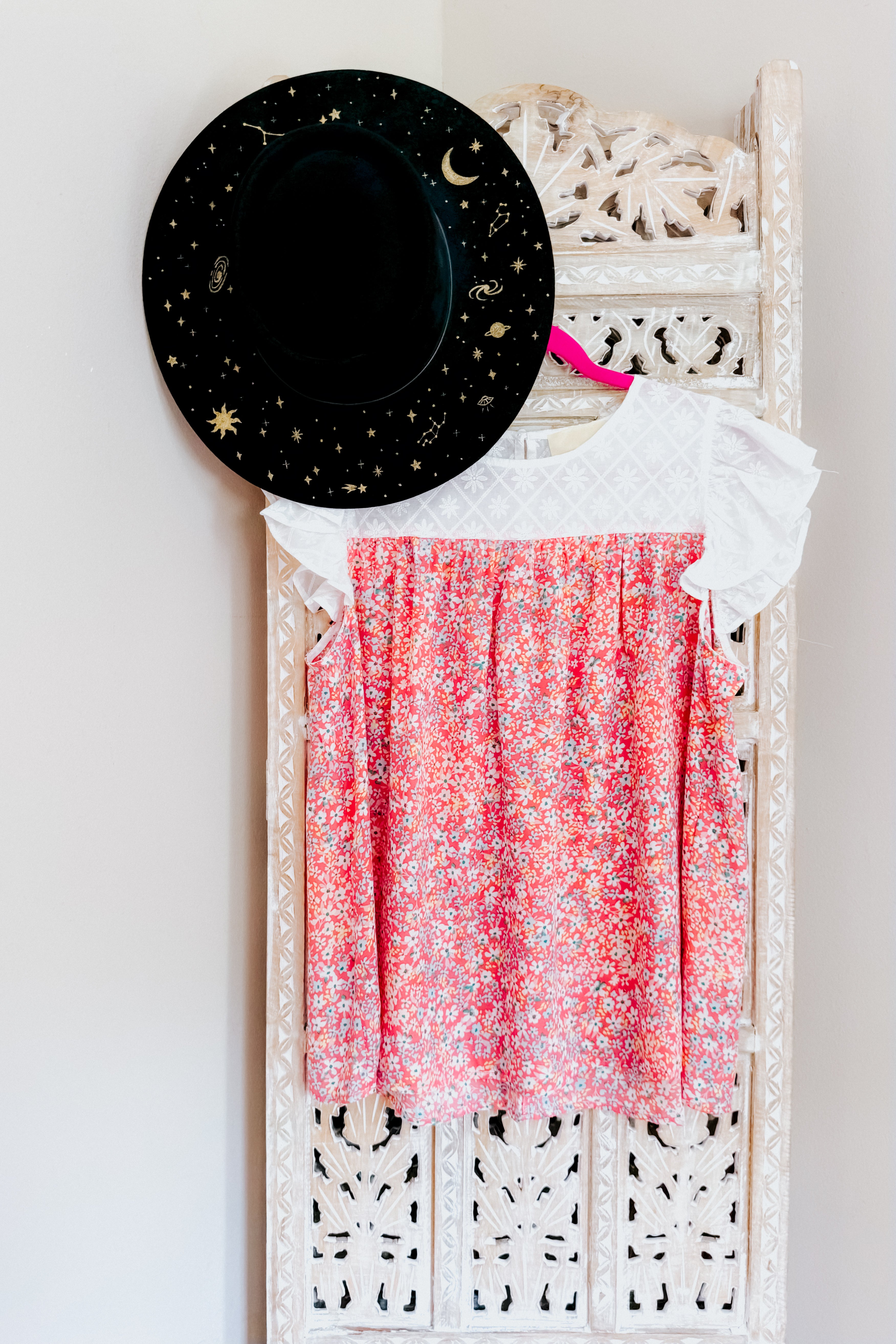 Pink floral dress with white textured yoke and flutter sleeves hanging on carved wooden screen with black starry hat