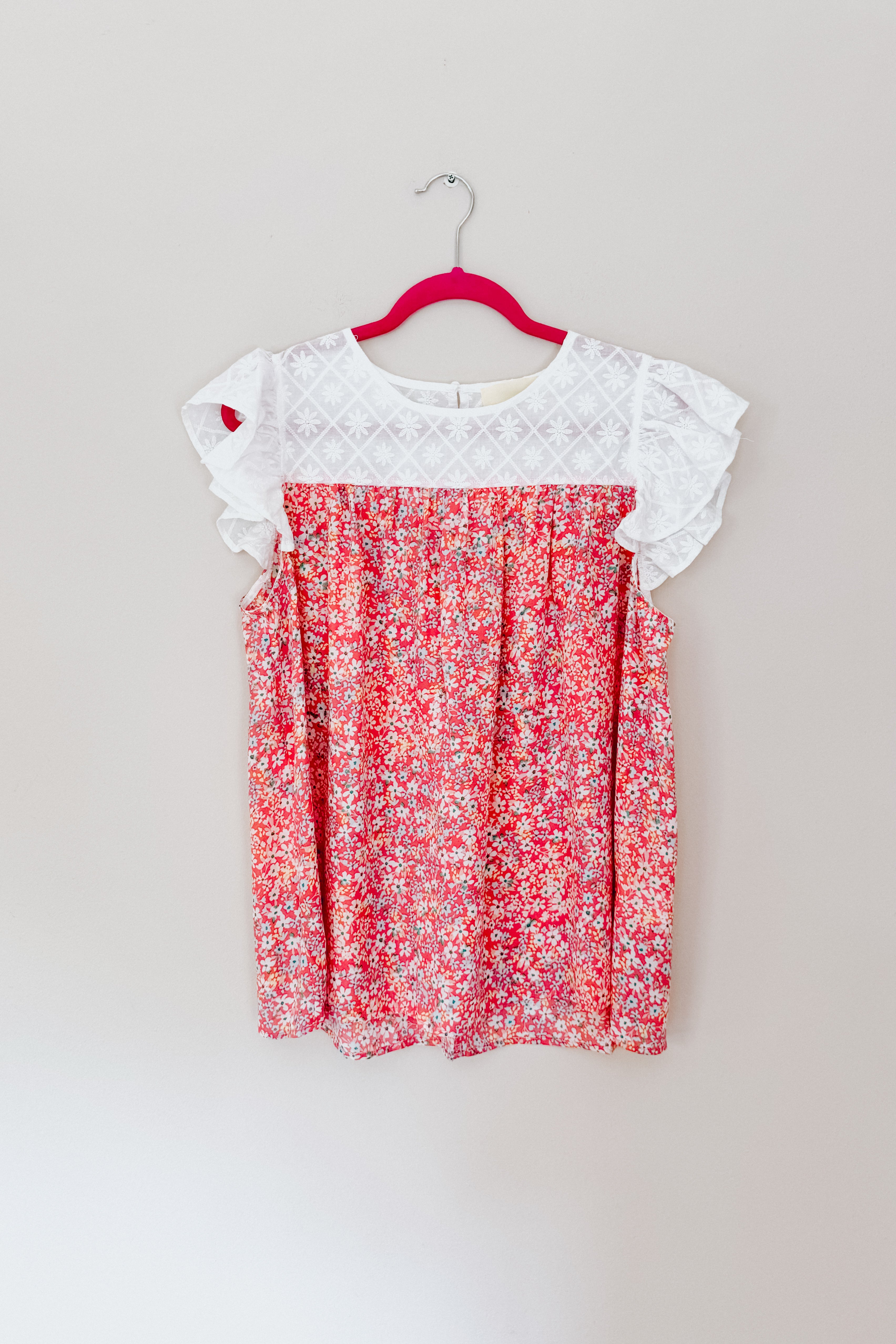 Red floral sleeveless blouse with white eyelet lace yoke and ruffled cap sleeves hanging on a pink hanger against a wall