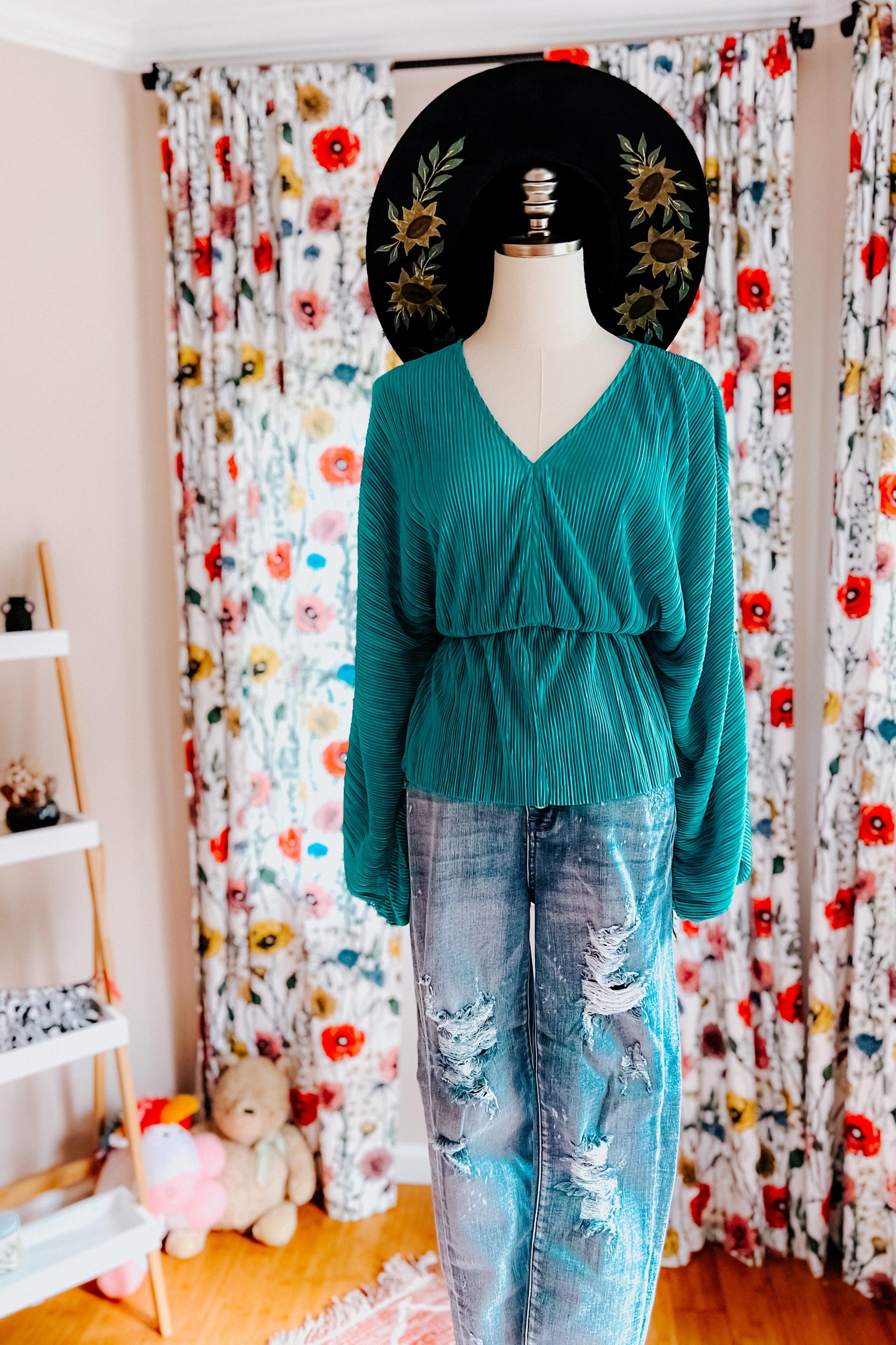 Teal pleated blouse with long sleeves paired with distressed light blue denim jeans on mannequin with floral backdrop