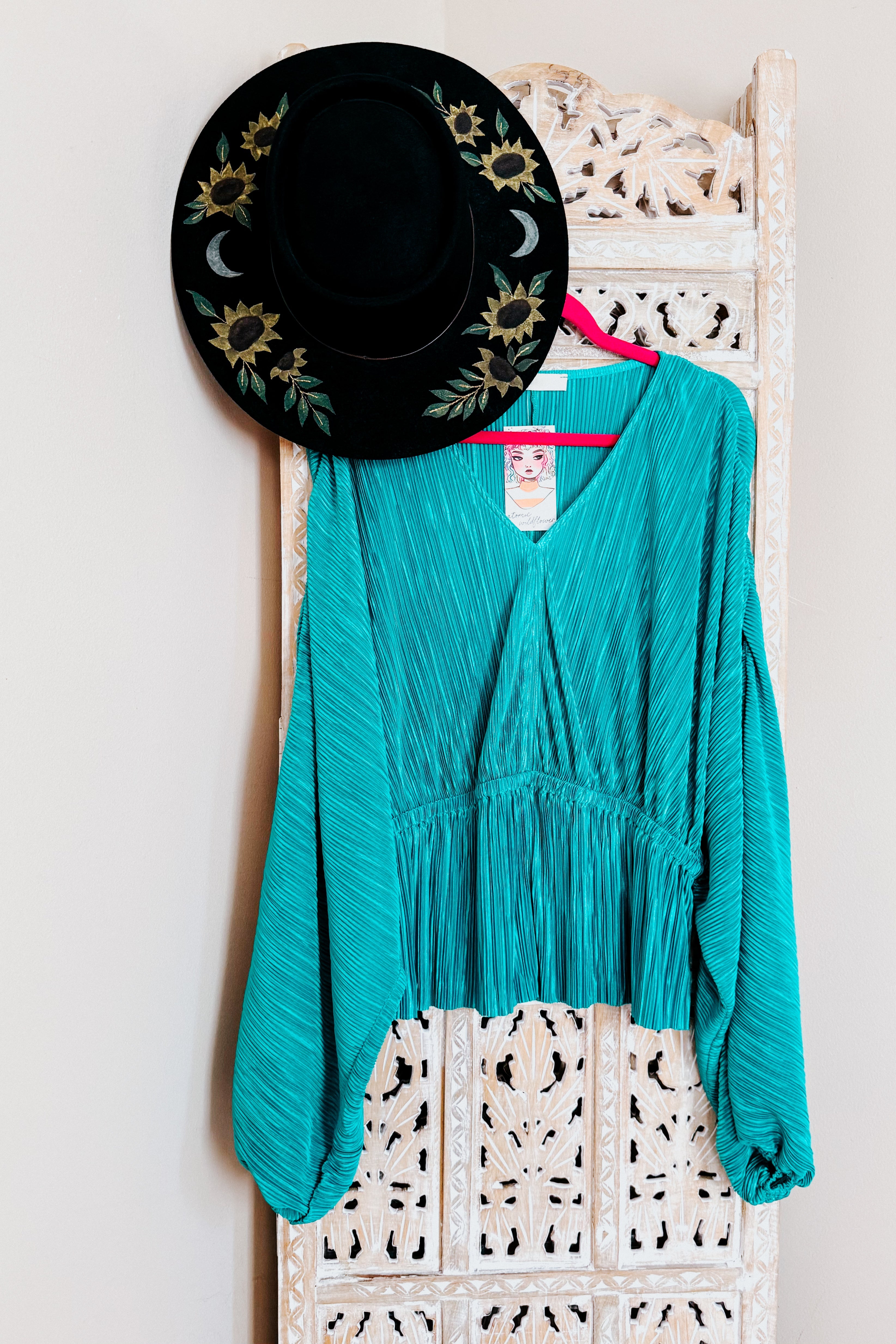 Teal textured blouse with voluminous sleeves on pink hanger paired with black wide-brimmed hat featuring sunflower embroidery