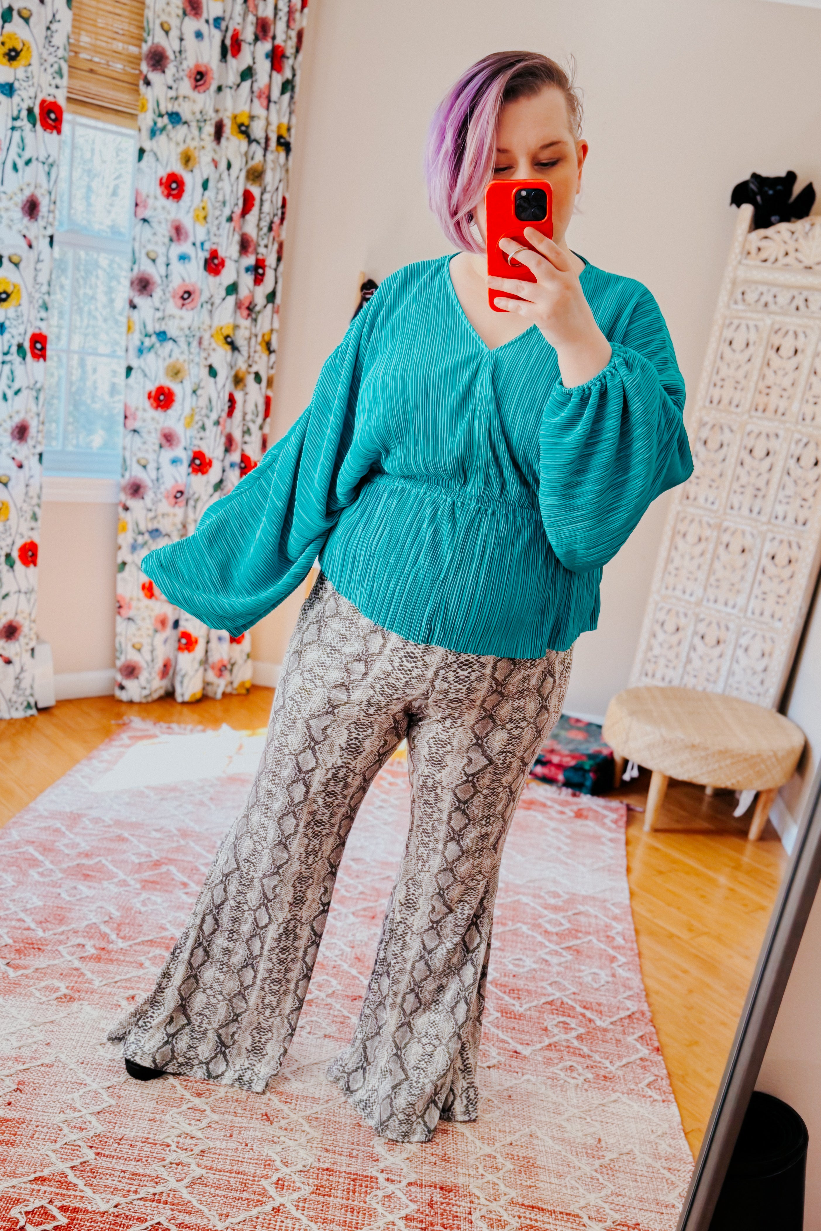 Woman with purple hair wearing teal textured blouse with wide sleeves and snake print flared pants taking mirror selfie indoors