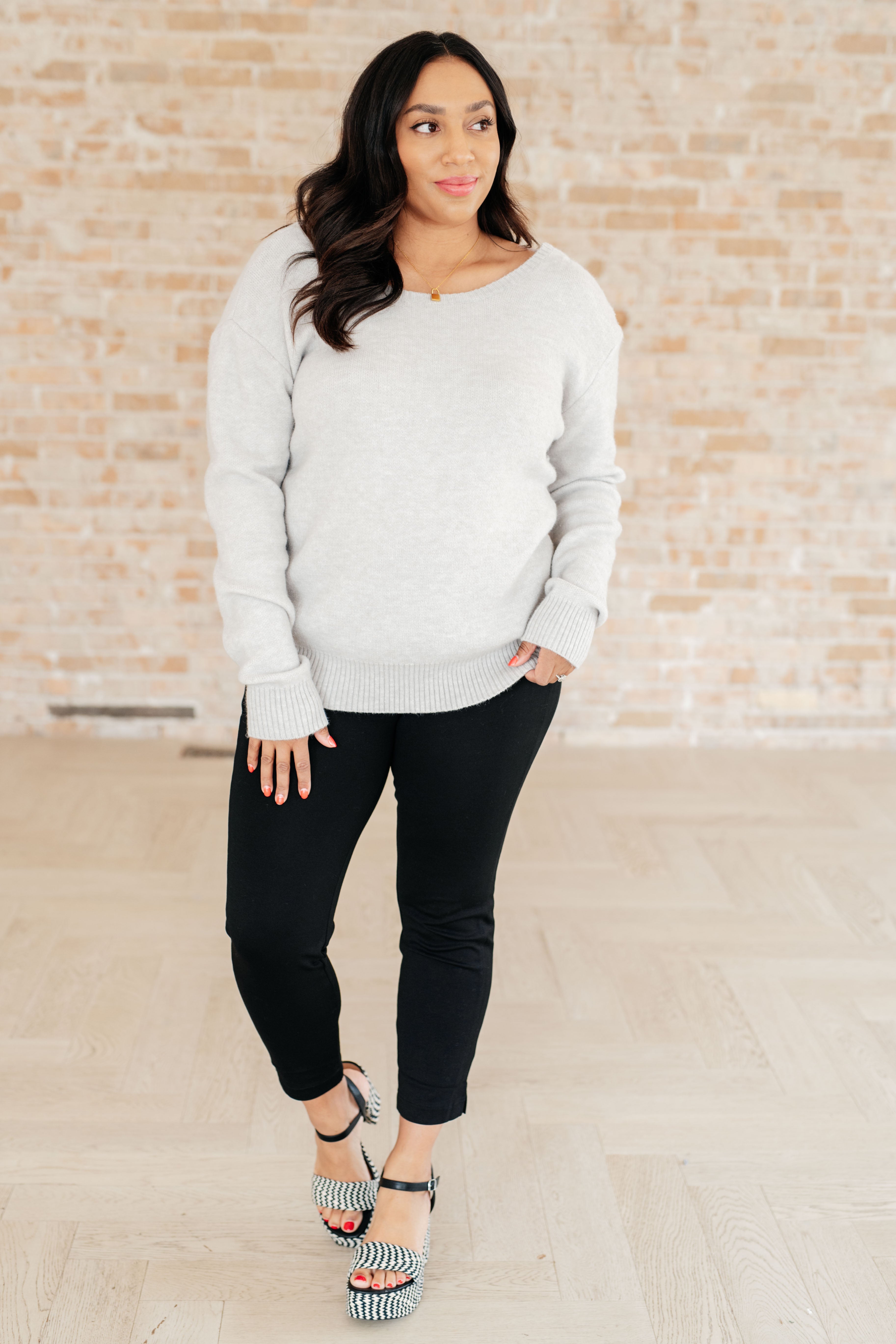Woman wearing light grey long-sleeve knit top with ribbed cuffs paired with fitted black pants and patterned heels indoors