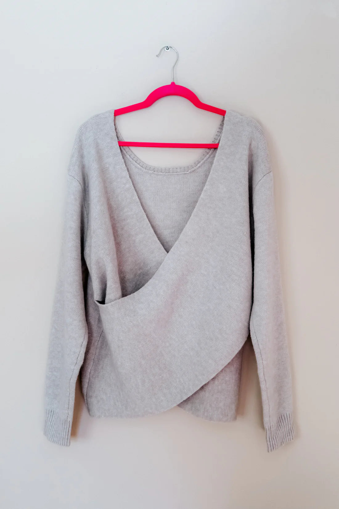 Grey knit sweater with an asymmetrical wrap front and ribbed cuffs hanging on a bright pink hanger against a white wall
