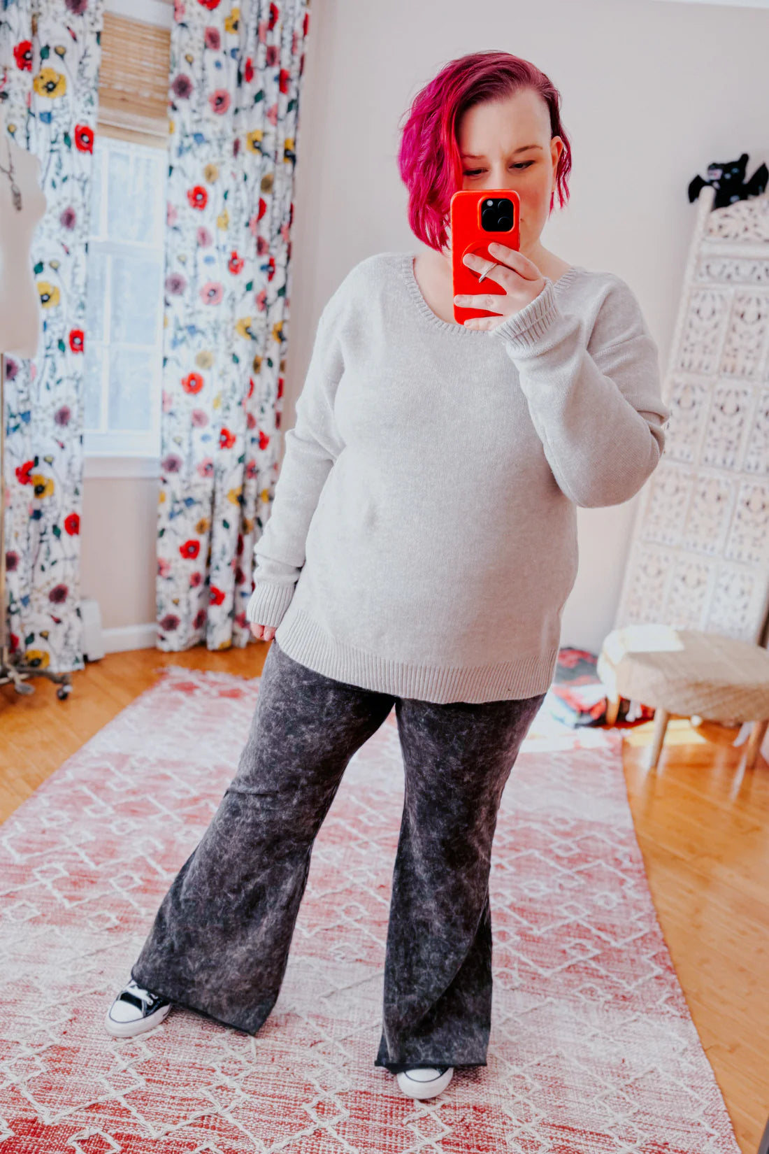 Woman with short magenta hair wearing a soft grey knit sweater and dark grey wide-leg pants taking a mirror selfie indoors