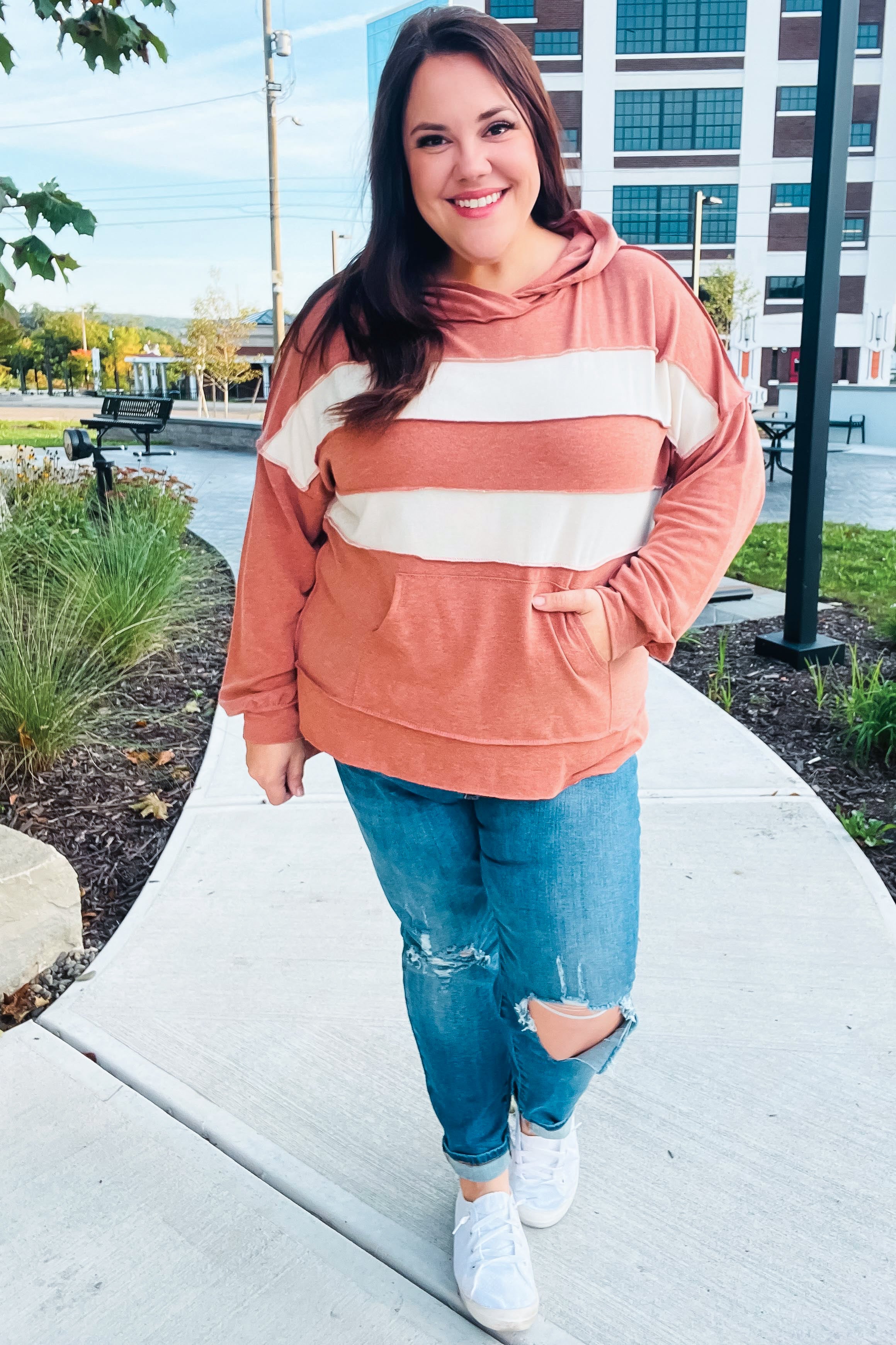 Smiling woman in a rust hoodie with broad cream horizontal stripes and ripped jeans walking on a sidewalk outdoors