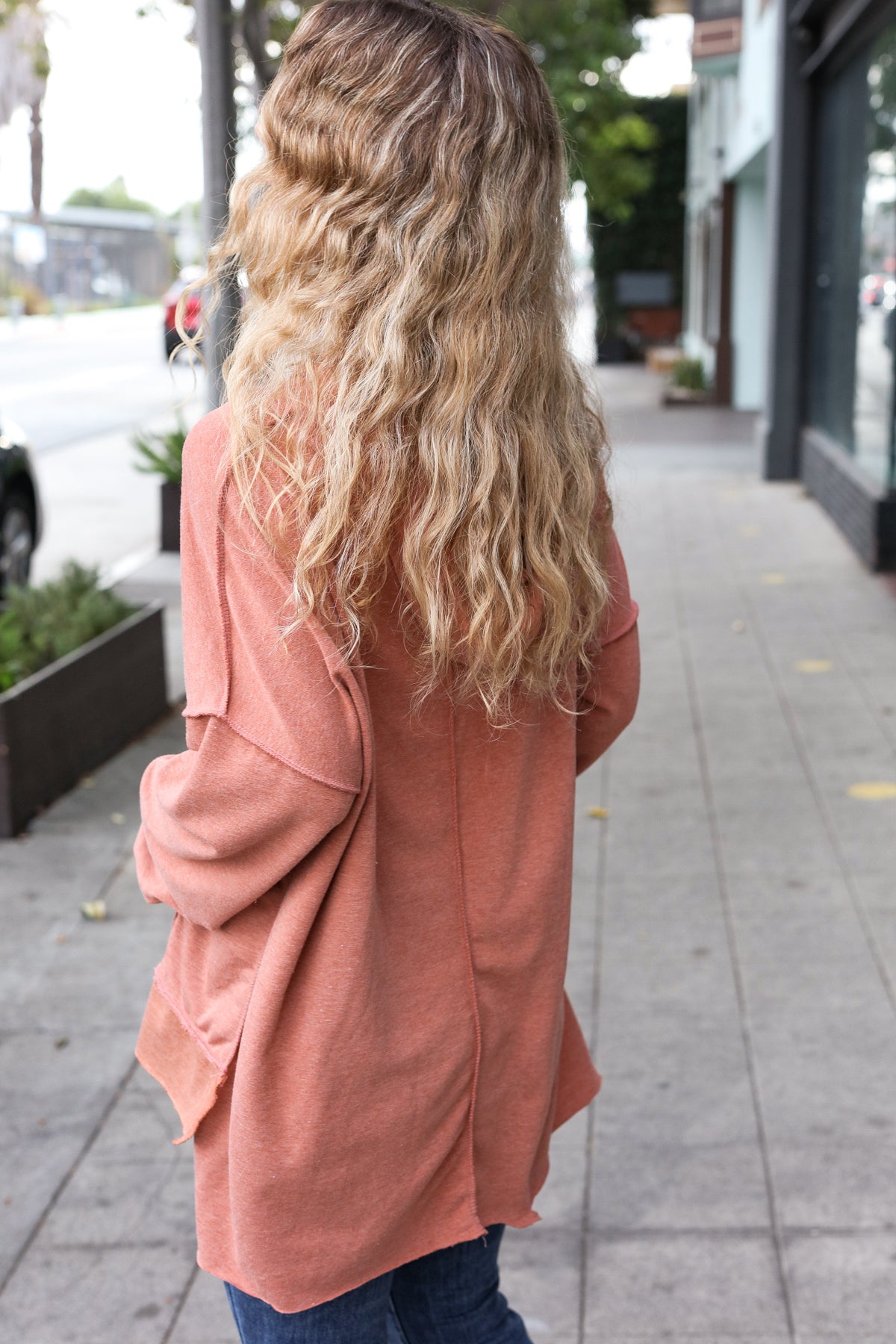 Woman with long wavy hair wearing a loose rust-colored sweater walking on an urban sidewalk outdoors