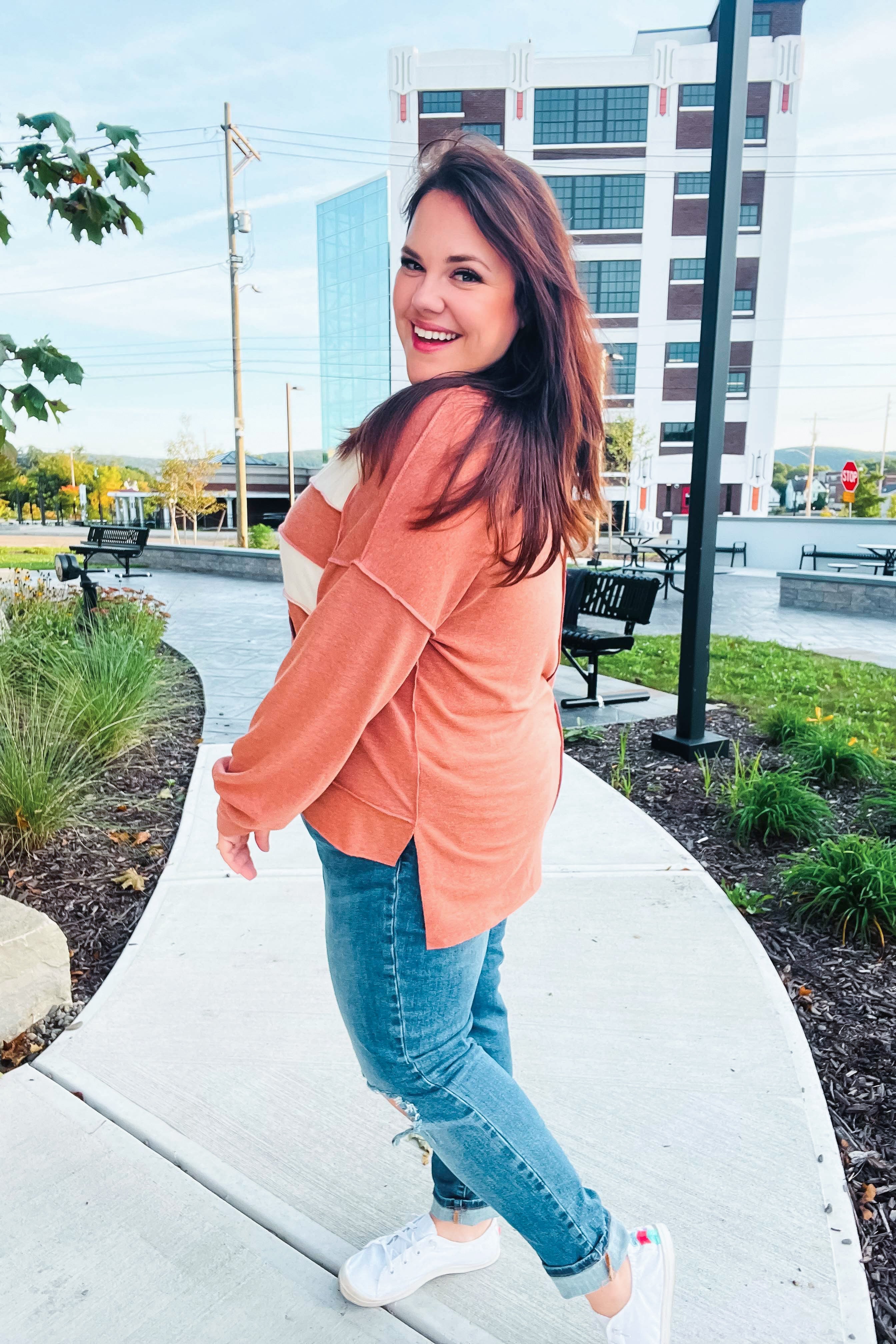 Happy woman with long brown hair wearing a rust-colored sweater with cream stripes and ripped jeans outdoors