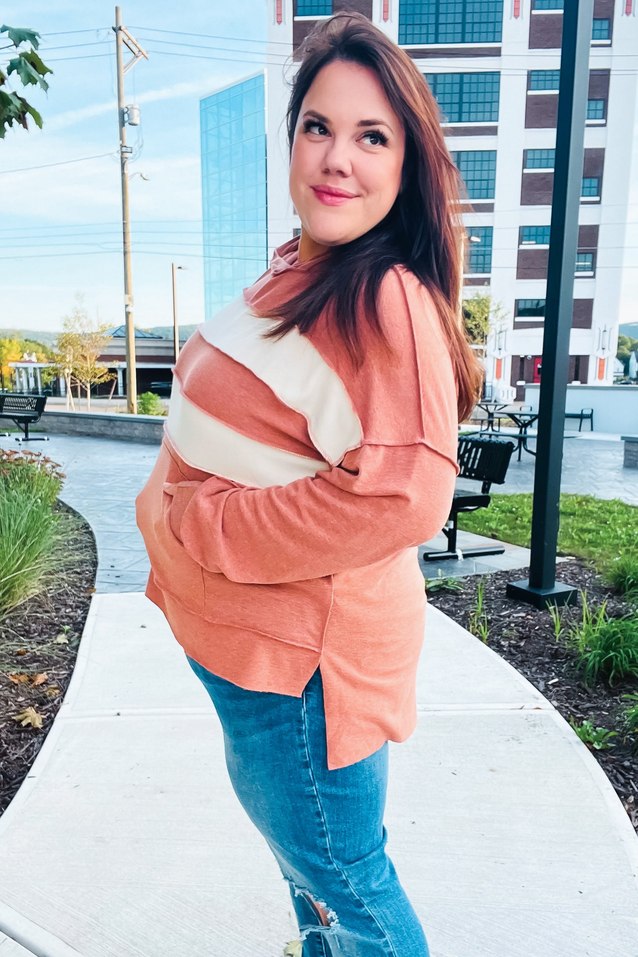 Woman wearing a rust-colored sweater with cream diagonal stripes and ripped blue jeans standing outdoors on a walkway