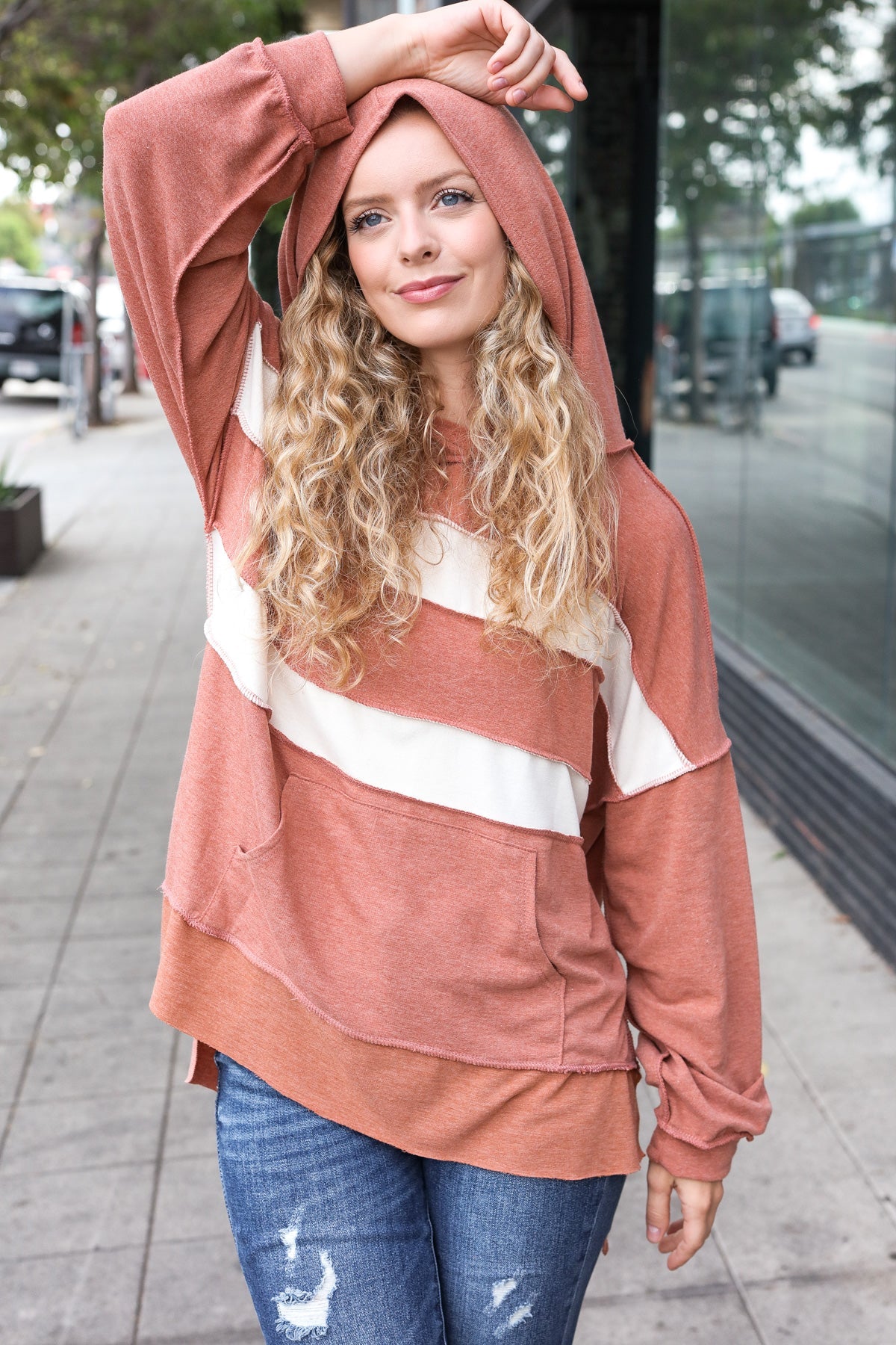 Blonde woman wearing a rust-colored hoodie with cream diagonal stripes and distressed blue jeans standing outdoors on sidewalk