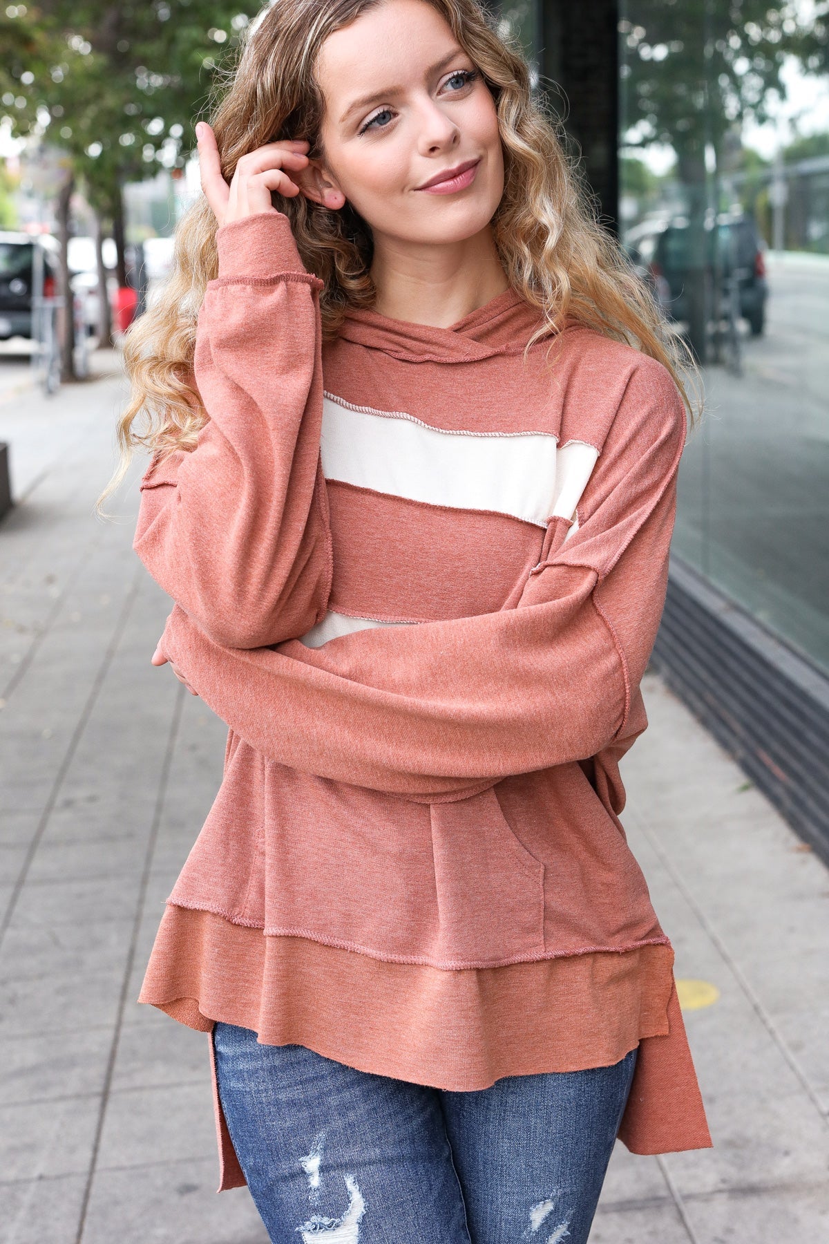 Young woman with curly hair wearing a rust-colored hoodie with a wide cream horizontal stripe outdoors