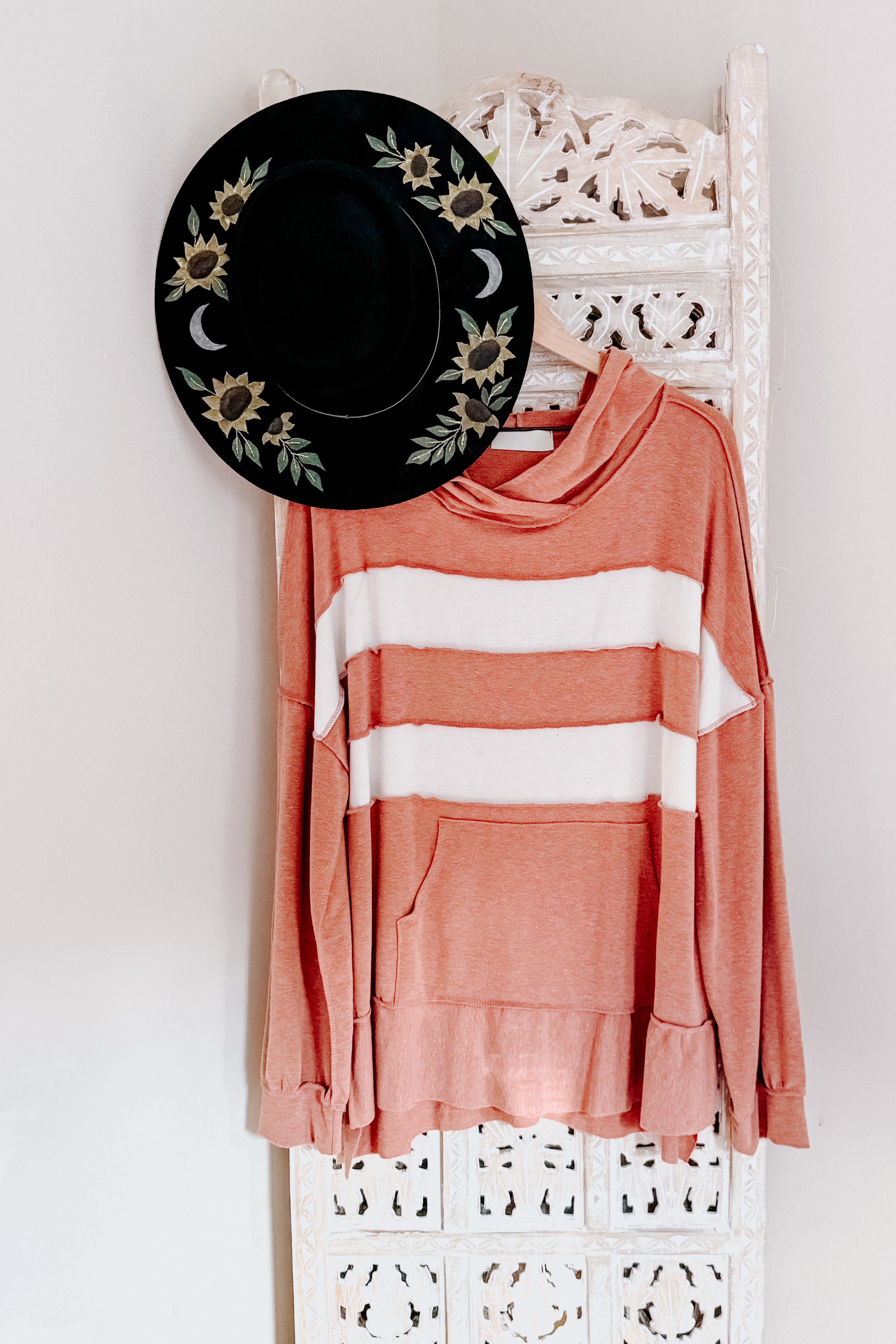 Rust-colored long sleeve top with two wide white horizontal stripes hanging beside a black hat with floral and moon designs